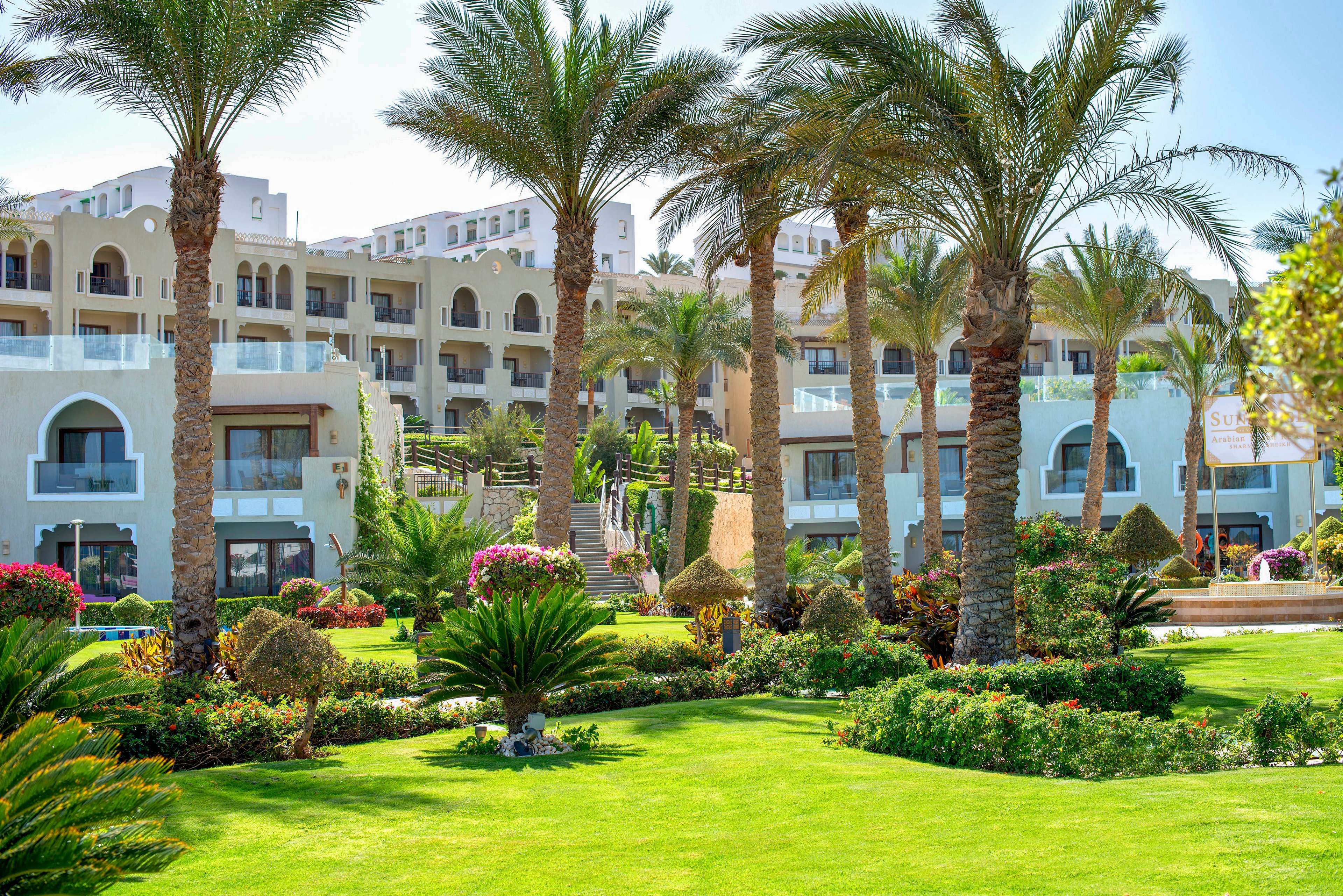 A large resort hotel complex rises behind a meticulously maintained green lawn and landscaped gardens. Tall palm trees and flowering shrubs frame the view, creating a tropical vacation atmosphere. The building features multiple balconies and arched windows, suggesting guest rooms overlooking the garden. Bright sunlight and clear skies enhance the feeling of a warm, relaxing holiday destination.