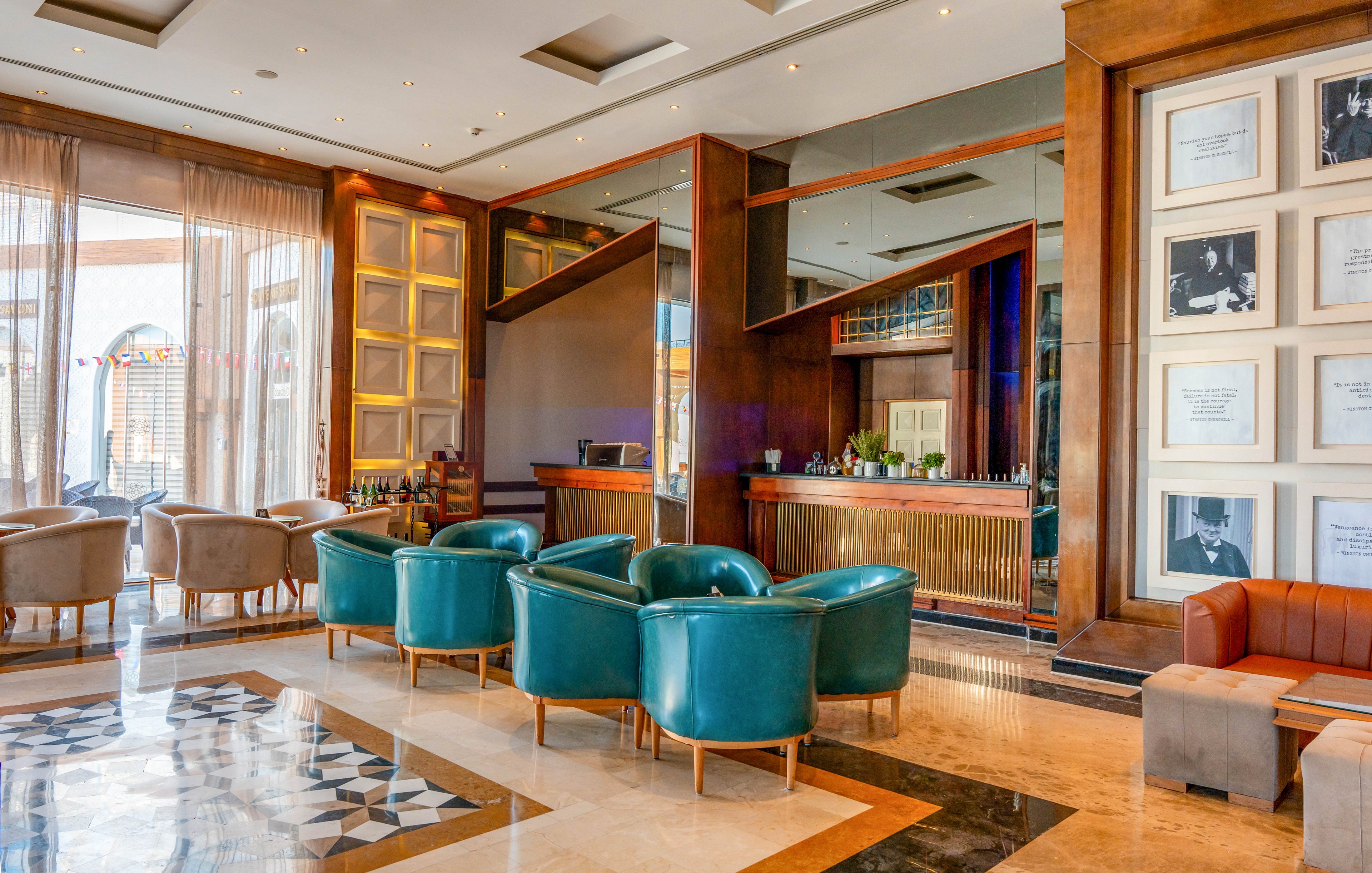 A stylish hotel lounge bar features teal leather armchairs arranged around small tables on a polished marble floor. The bar counter with bottles and glassware sits against a mirrored wall framed by warm wooden panels. Large windows with sheer curtains allow natural light to brighten the interior and highlight geometric floor patterns. Framed black and white photographs line one wall, adding a sophisticated, gallery-like atmosphere. The space appears calm and inviting, suitable for relaxed socializing or quiet conversation.