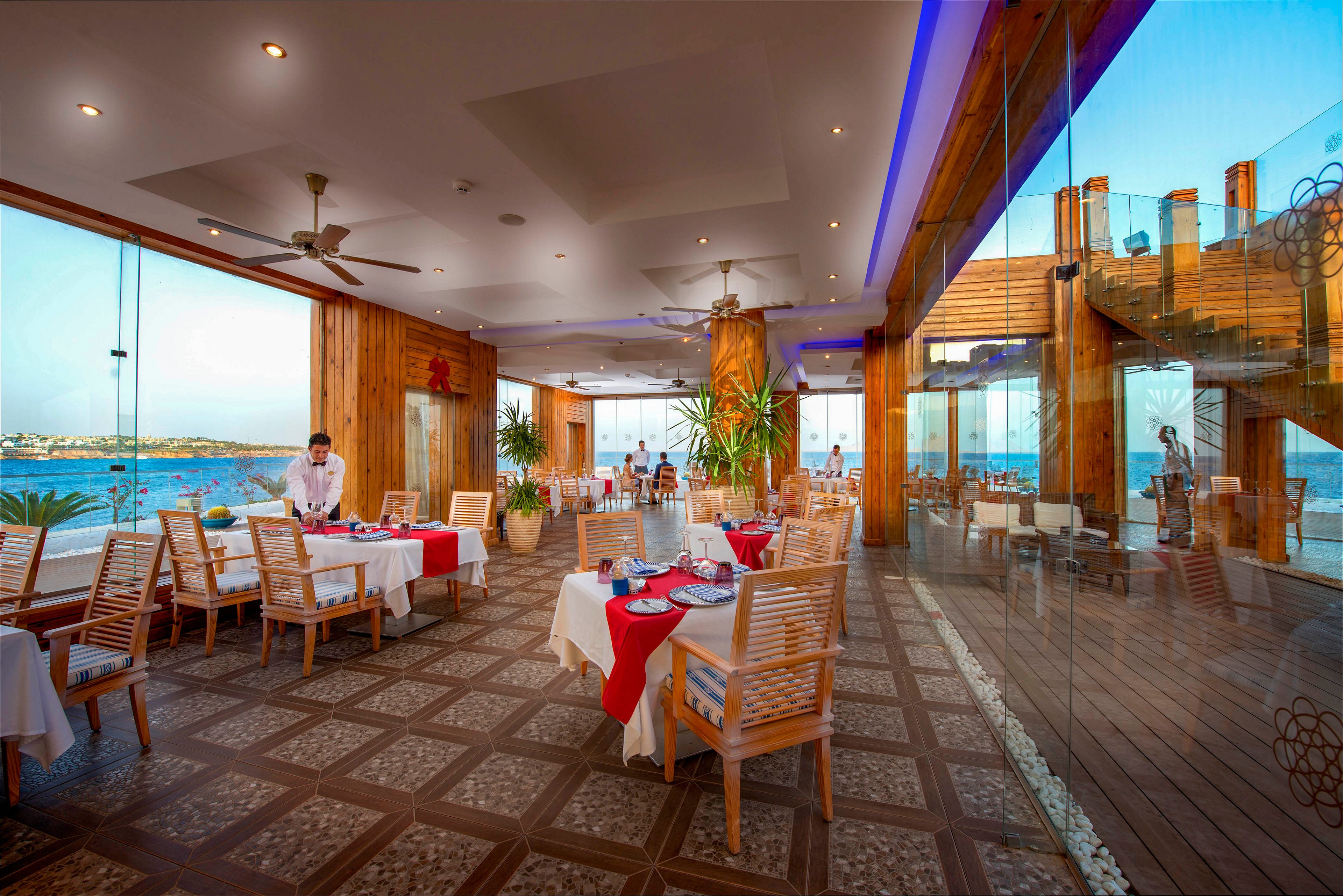 A spacious seaside restaurant features wooden columns, tiled floors, and large glass walls overlooking the water. Tables are set with white cloths and red accents, while a waiter prepares the dining area. Warm interior lighting contrasts with the blue tones of the sea and sky outside. Reflections in the glass emphasize the open, airy atmosphere and coastal setting.