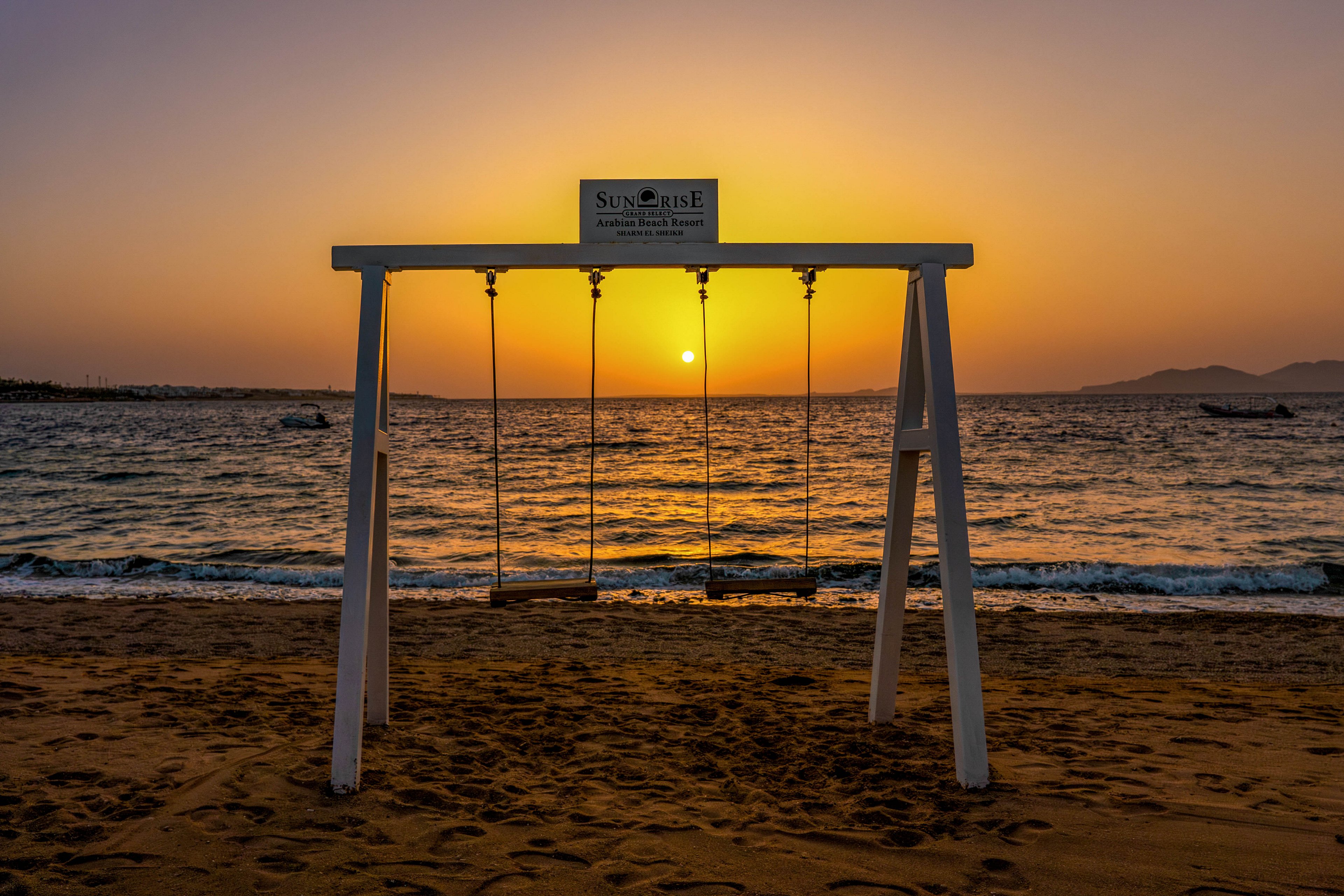 A set of wooden swings hangs from a white frame on a sandy beach at sunset. The sun is perfectly aligned between the swings, casting a warm golden glow over the calm sea. Gentle waves roll toward the shore while distant boats are visible on the horizon. The sky transitions from deep orange near the sun to darker tones above, creating a peaceful and romantic coastal scene.