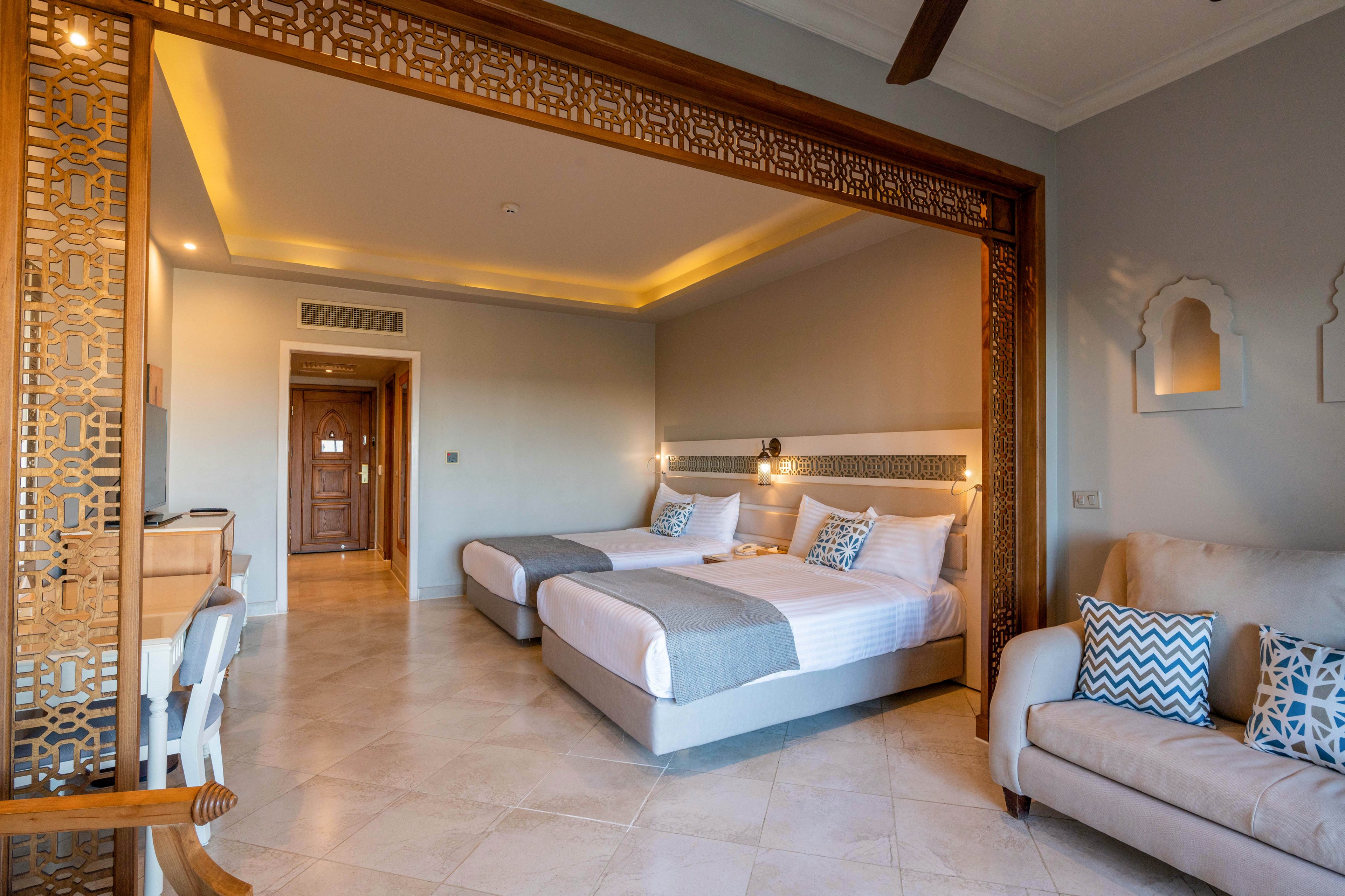 A spacious hotel room features two neatly made twin beds with gray runners and patterned pillows. The room is divided by an ornate wooden screen that frames the sleeping area. Soft, warm lighting from the ceiling and wall lamps creates a cozy atmosphere. A beige sofa with blue and white cushions sits to the side, enhancing the comfortable, contemporary design.