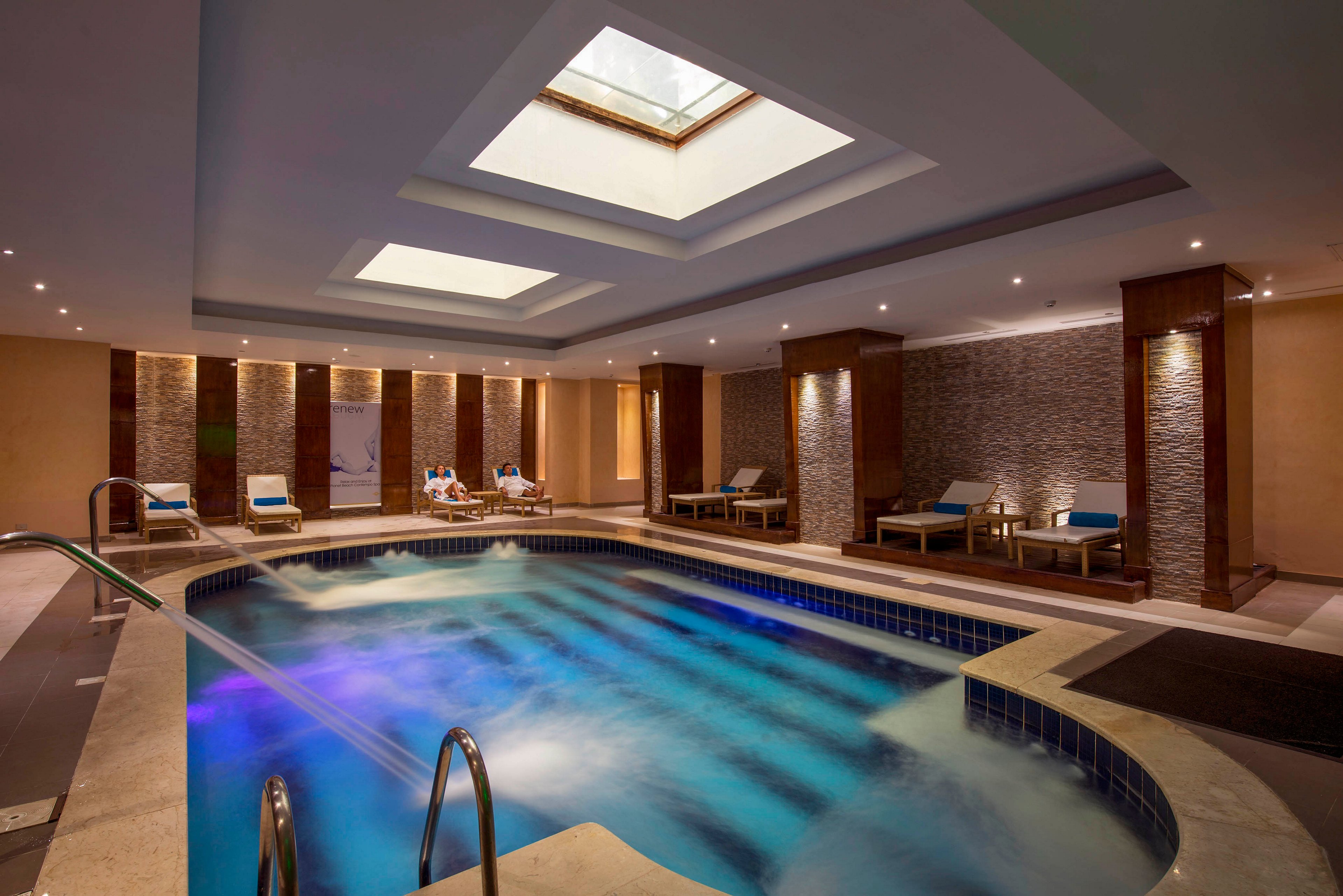 A modern indoor spa pool glows with blue and purple lighting beneath a ceiling skylight. Comfortable lounge chairs line the sides of the pool, with a few people relaxing in the background. Warm recessed lighting and textured stone walls create a tranquil, upscale atmosphere. Stainless steel handrails and bubbling water emphasize the wellness and relaxation setting.