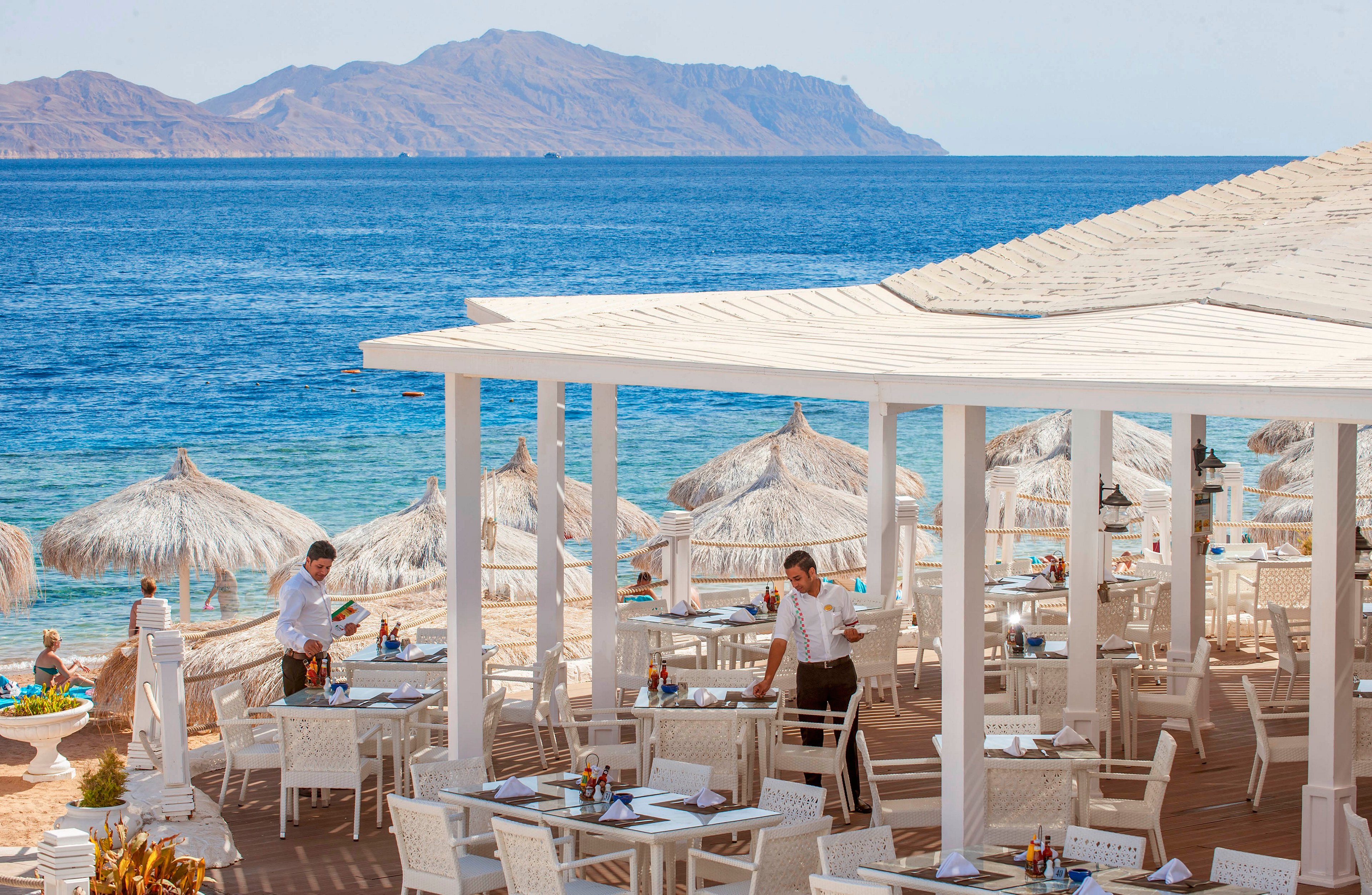 An open-air restaurant terrace overlooks a bright blue sea and distant rocky islands. White tables and chairs are arranged neatly under a pergola, with waitstaff preparing the dining area. Straw parasols line the sandy beach below, creating a relaxed coastal resort atmosphere. The scene suggests a sunny vacation setting ideal for seaside dining and leisure.