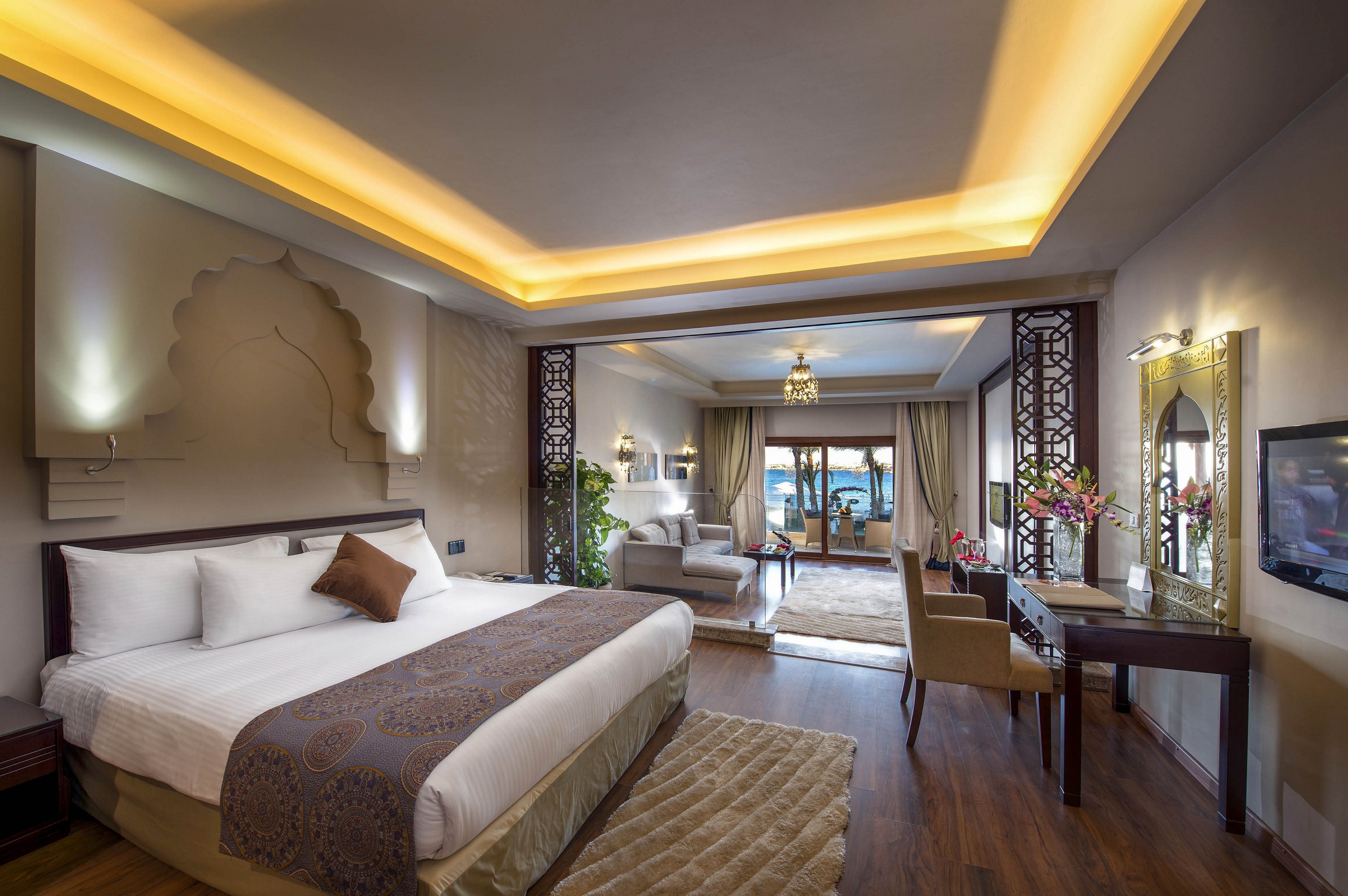 A spacious luxury hotel suite features a large bed with decorative pillows and a patterned runner. Warm recessed ceiling lighting and wooden floors create an inviting atmosphere. A sitting area with sofas and a desk leads toward large glass doors opening to a balcony with a view of the sea and palm trees. Elegant decor elements, including wall art, mirrors, and fresh flowers, enhance the upscale resort ambiance.