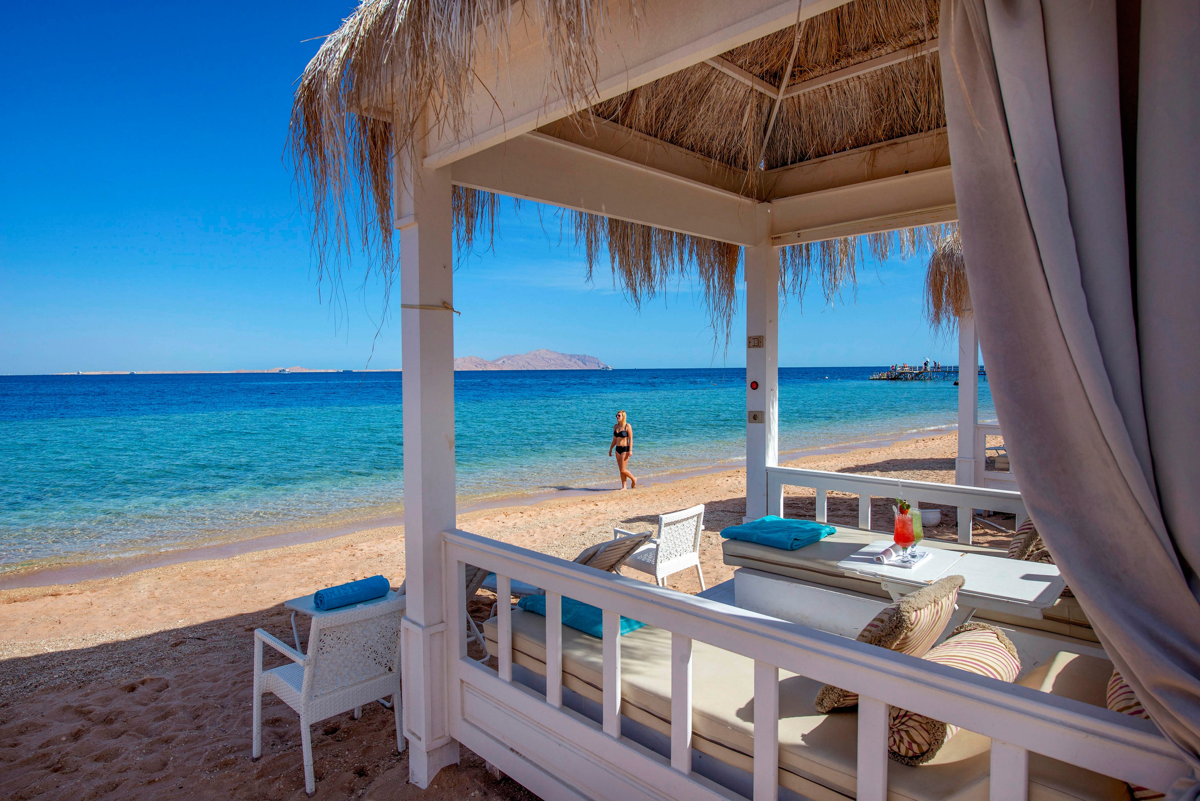 A luxurious white cabana with cushioned seating sits on a sandy beach overlooking a calm turquoise sea. A person in swimwear walks along the shoreline in the background under a clear blue sky. The cabana features straw roofing, draped curtains, and a table set with drinks and towels, suggesting a relaxing resort setting. Bright sunlight and vivid colors create a serene, vacation atmosphere.
