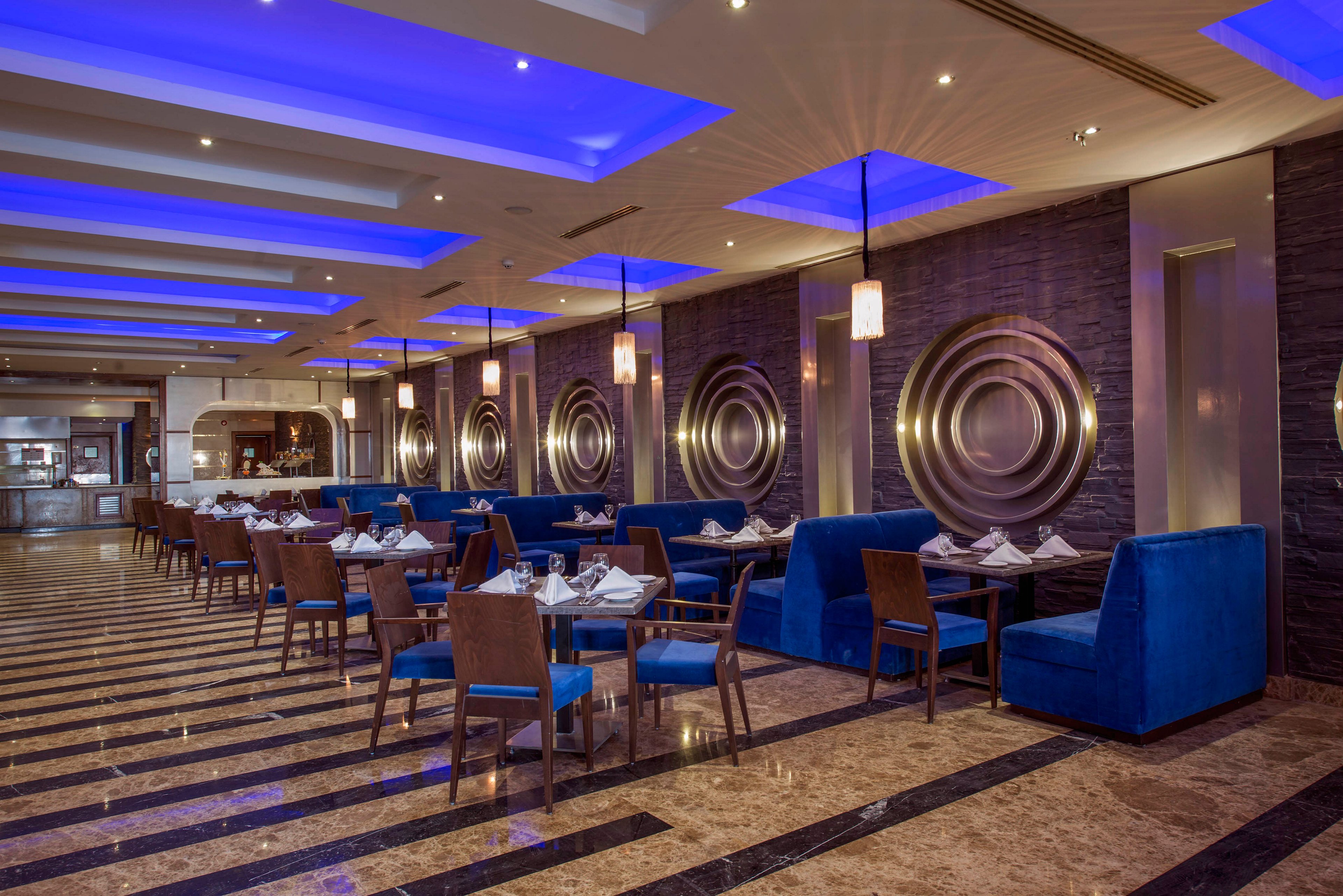 A contemporary restaurant dining area features rows of wooden tables set with white napkins and glassware. Plush blue banquettes line the walls, complemented by matching blue ceiling lights that create a dramatic ambiance. Circular metallic wall accents add a stylish focal point along the length of the room. The polished marble floor with dark striped inlays enhances the upscale, modern atmosphere.