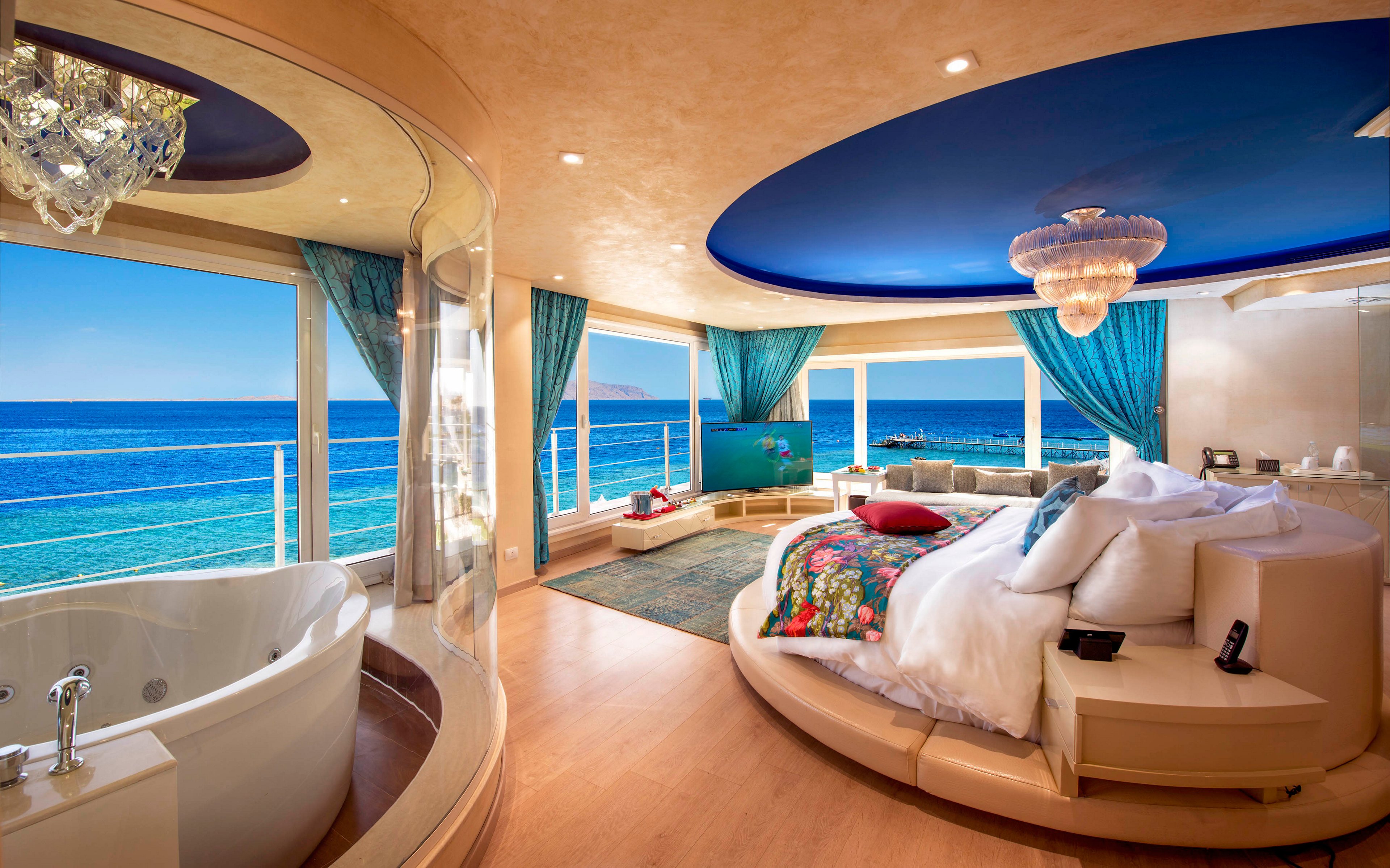 A luxurious circular bed sits in a spacious oceanfront bedroom suite with panoramic floor-to-ceiling windows. The room features a large in-room jacuzzi tub near the windows, offering views of the bright blue sea and clear sky. Rich turquoise curtains, a blue ceiling dome, and crystal chandeliers create an opulent, resort-like atmosphere. Warm wood flooring and soft lighting enhance the relaxing, romantic mood of the coastal retreat.