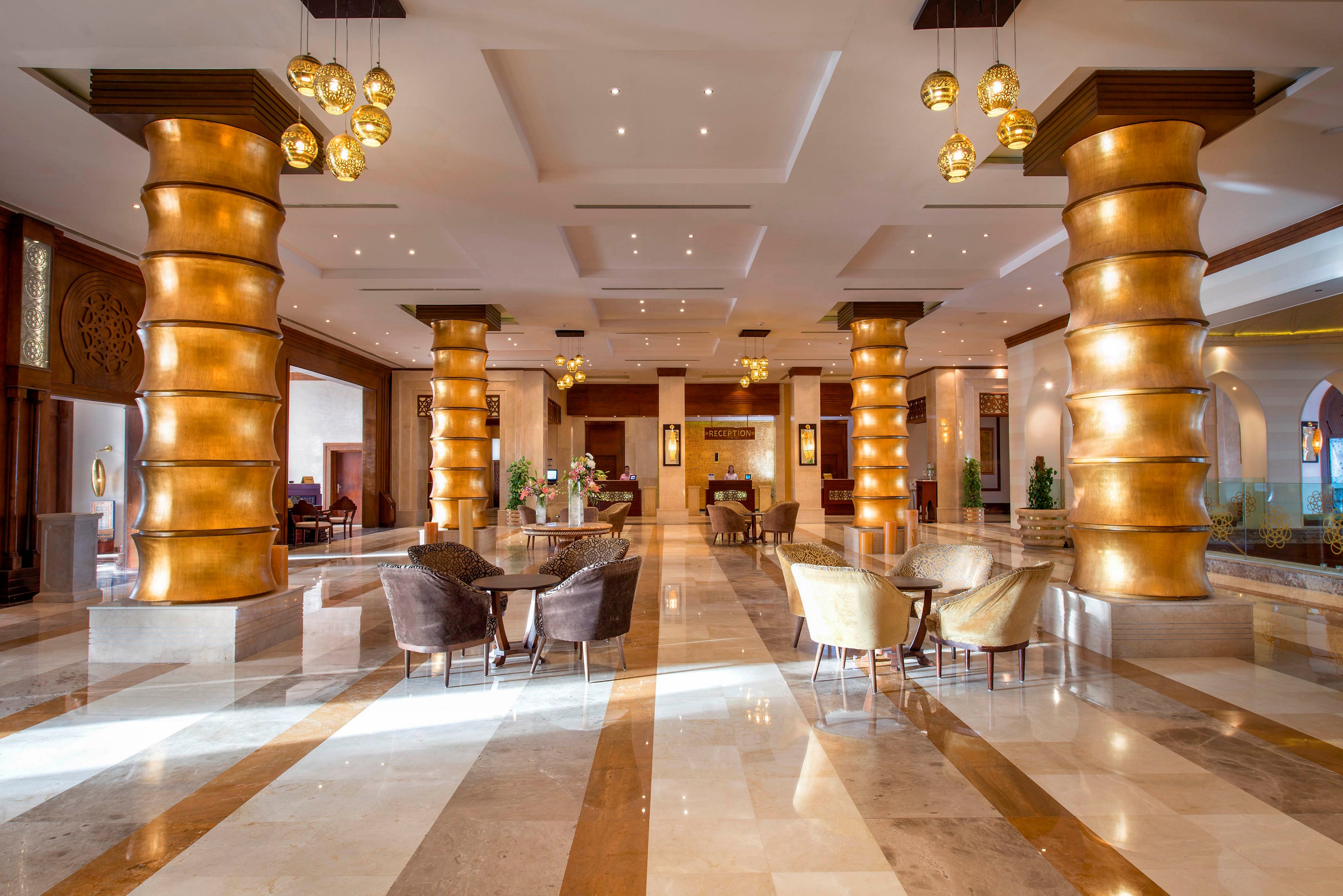 A spacious hotel lobby features striking golden cylindrical columns arranged in two rows. Polished marble floors and a high white ceiling with recessed lighting create a bright, elegant atmosphere. Clusters of armchairs and small tables provide seating areas along the central walkway. Warm ambient lighting from hanging lamps and wall fixtures enhances the sophisticated, welcoming feel of the interior.