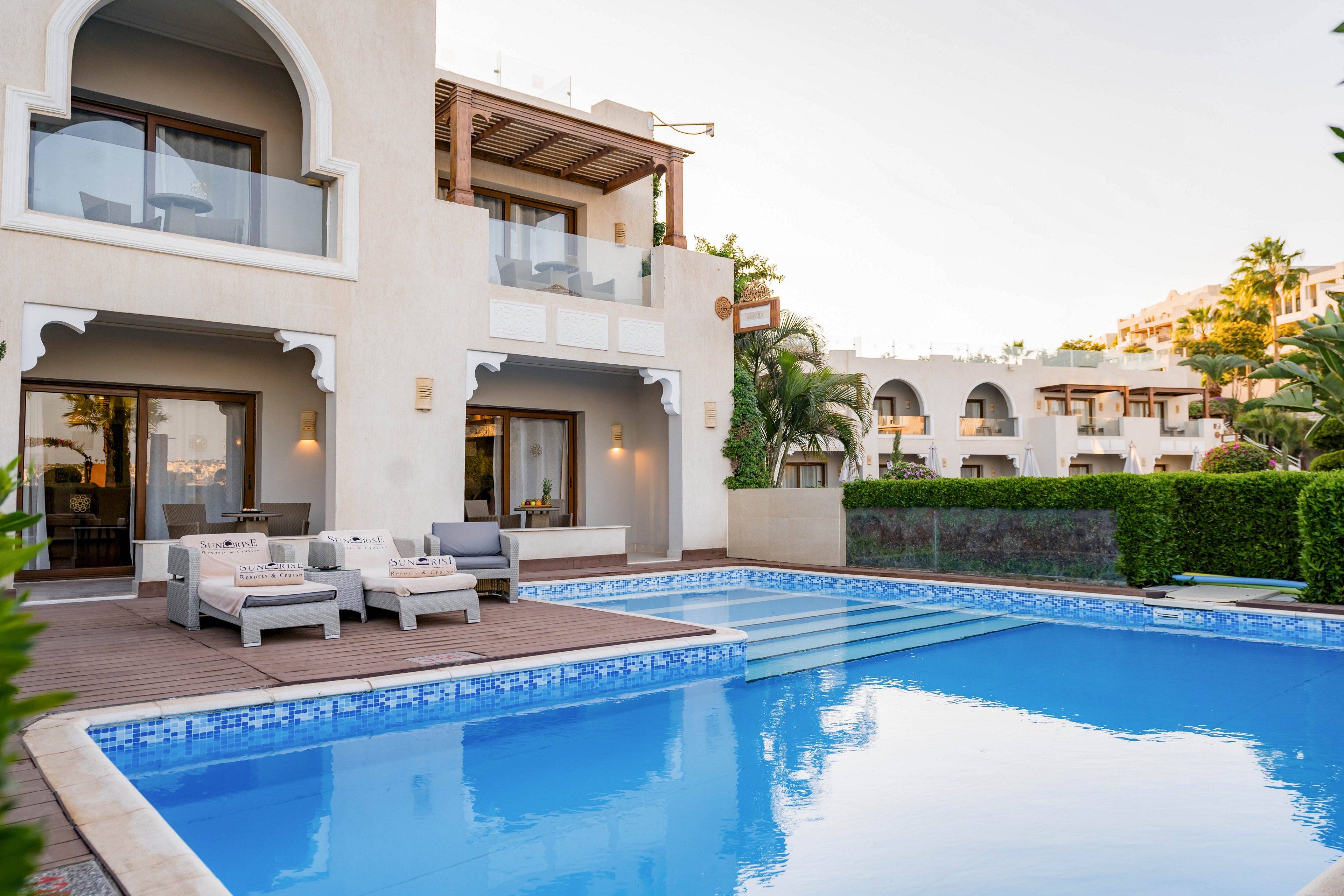 A modern luxury villa overlooks a clear blue swimming pool in a resort setting. The building features arched balconies, white walls, and large glass doors opening onto a wooden deck. Outdoor lounge furniture is arranged beside the pool, creating a comfortable relaxation area. Manicured hedges, palm trees, and neighboring villas enhance the upscale, tranquil atmosphere.