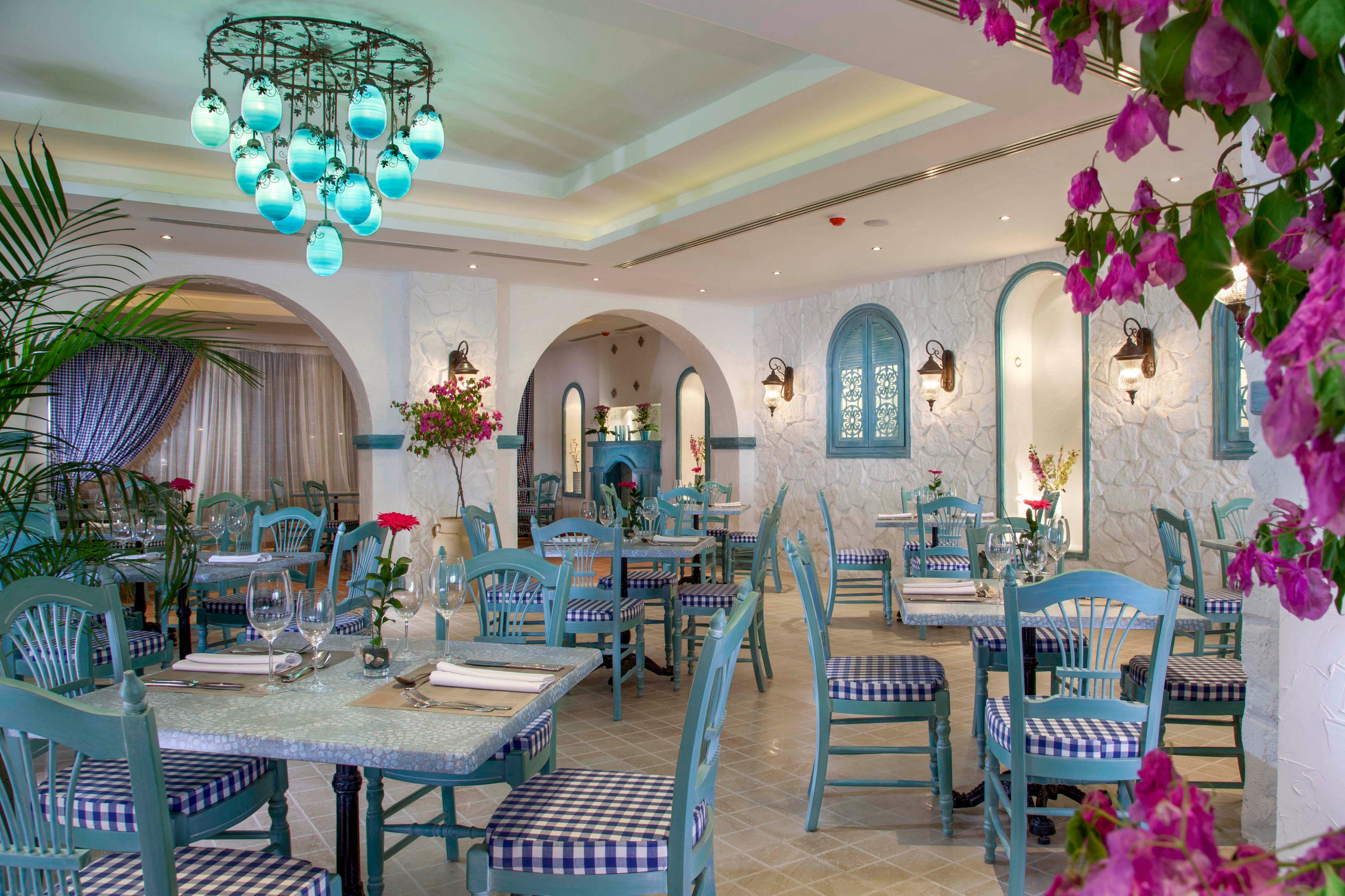 A bright coastal-style restaurant interior features turquoise wooden chairs and tables arranged neatly around the room. The space is decorated with checkered seat cushions, floral centerpieces, and hanging pendant lights with blue glass shades. Arched windows and wall niches add Mediterranean character, while pink flowers frame the foreground. The overall atmosphere feels airy, inviting, and ready for guests.