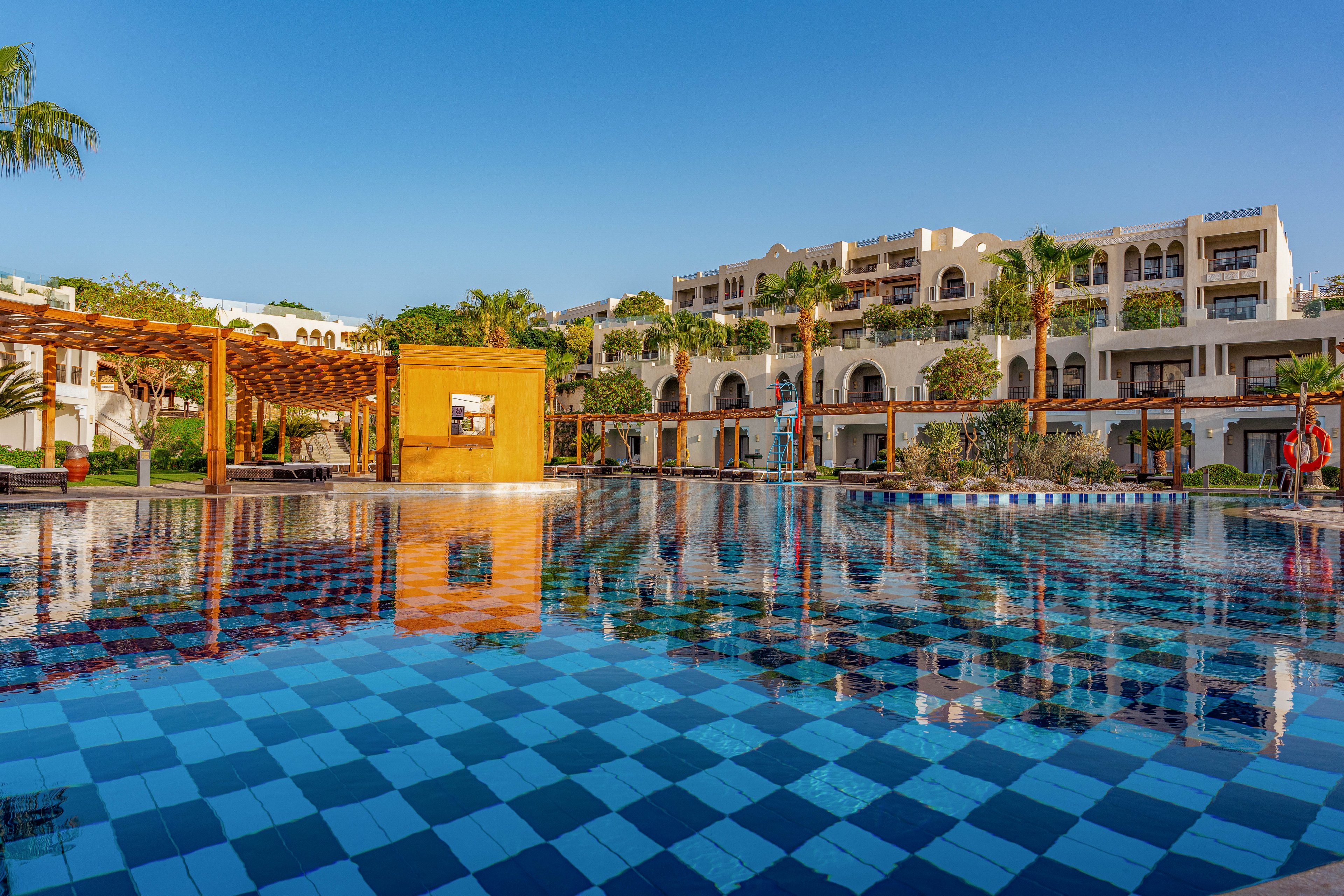 A large resort swimming pool with blue checkered tiles reflects the surrounding hotel buildings. The scene is set on a clear, sunny day with a bright blue sky. Palm trees, shaded pergolas, and balconies create a relaxed vacation atmosphere. The calm water and warm light suggest a tranquil, upscale holiday destination.