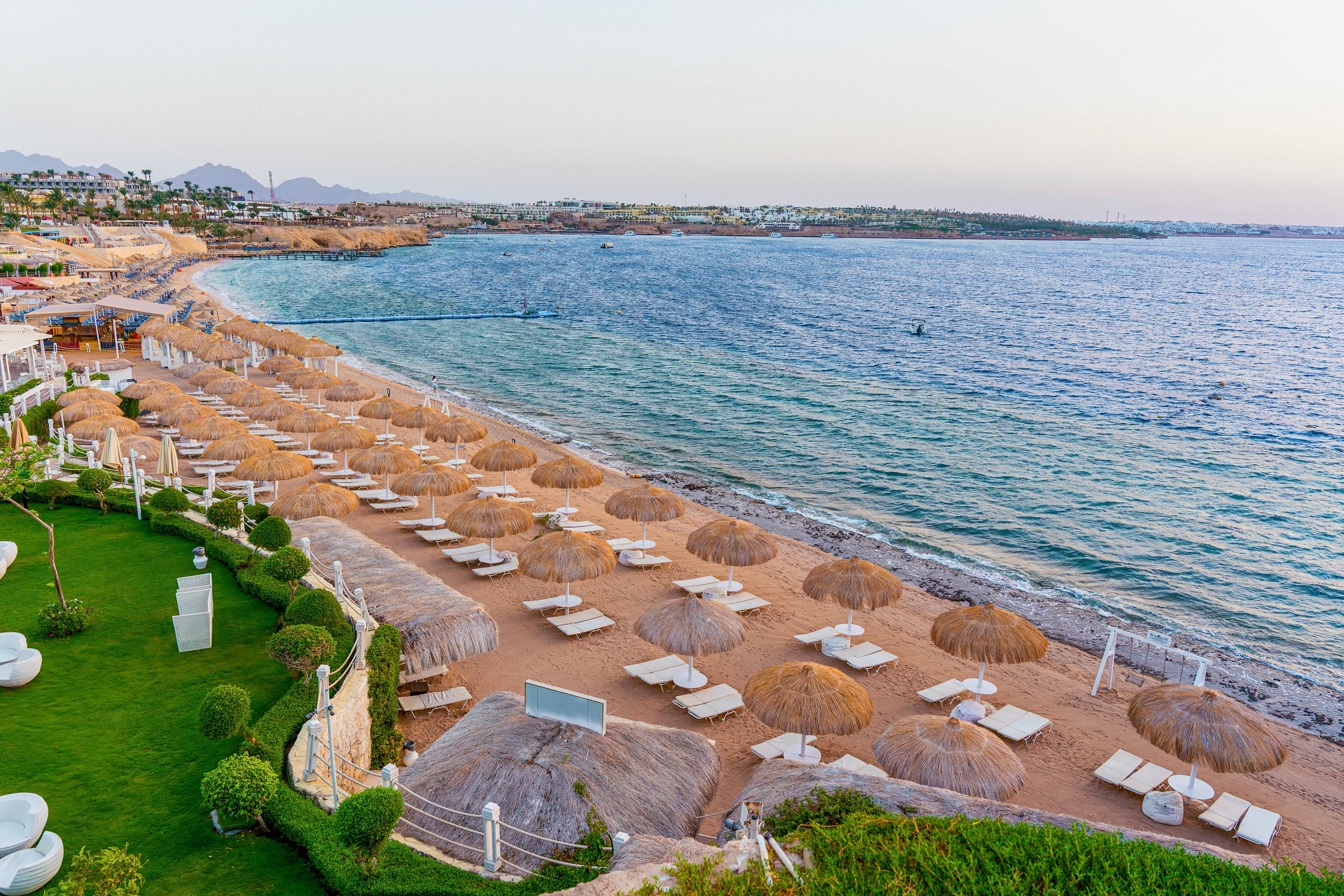 A tranquil resort beach is lined with rows of straw umbrellas and white sun loungers facing a calm sea. The shoreline curves gently, with clear blue water stretching to the horizon under a soft evening or morning sky. On the left, manicured green lawns and landscaped gardens border the sandy beach, adding contrast to the coastal scene. The overall atmosphere is peaceful and inviting, suggesting a relaxing holiday destination.