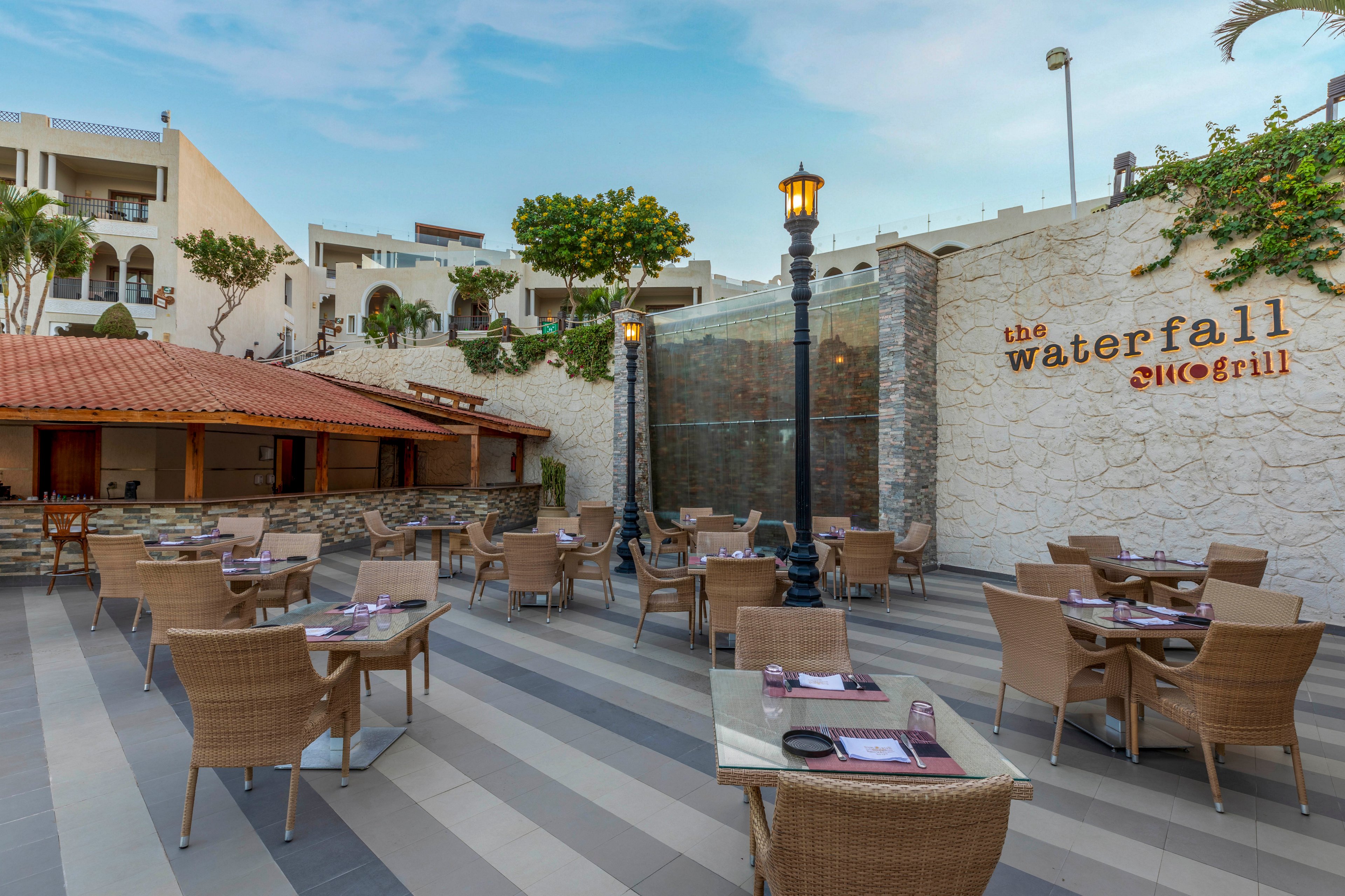 An open-air restaurant terrace features wicker chairs and glass-topped tables neatly arranged for guests. The setting is a modern resort courtyard with striped flooring and a central glass waterfall structure. Warm lamppost lighting and a red-tiled roof bar area add to the inviting atmosphere. The words "the waterfall" and "grill" are visible on the stone wall, emphasizing the dining venue’s name.