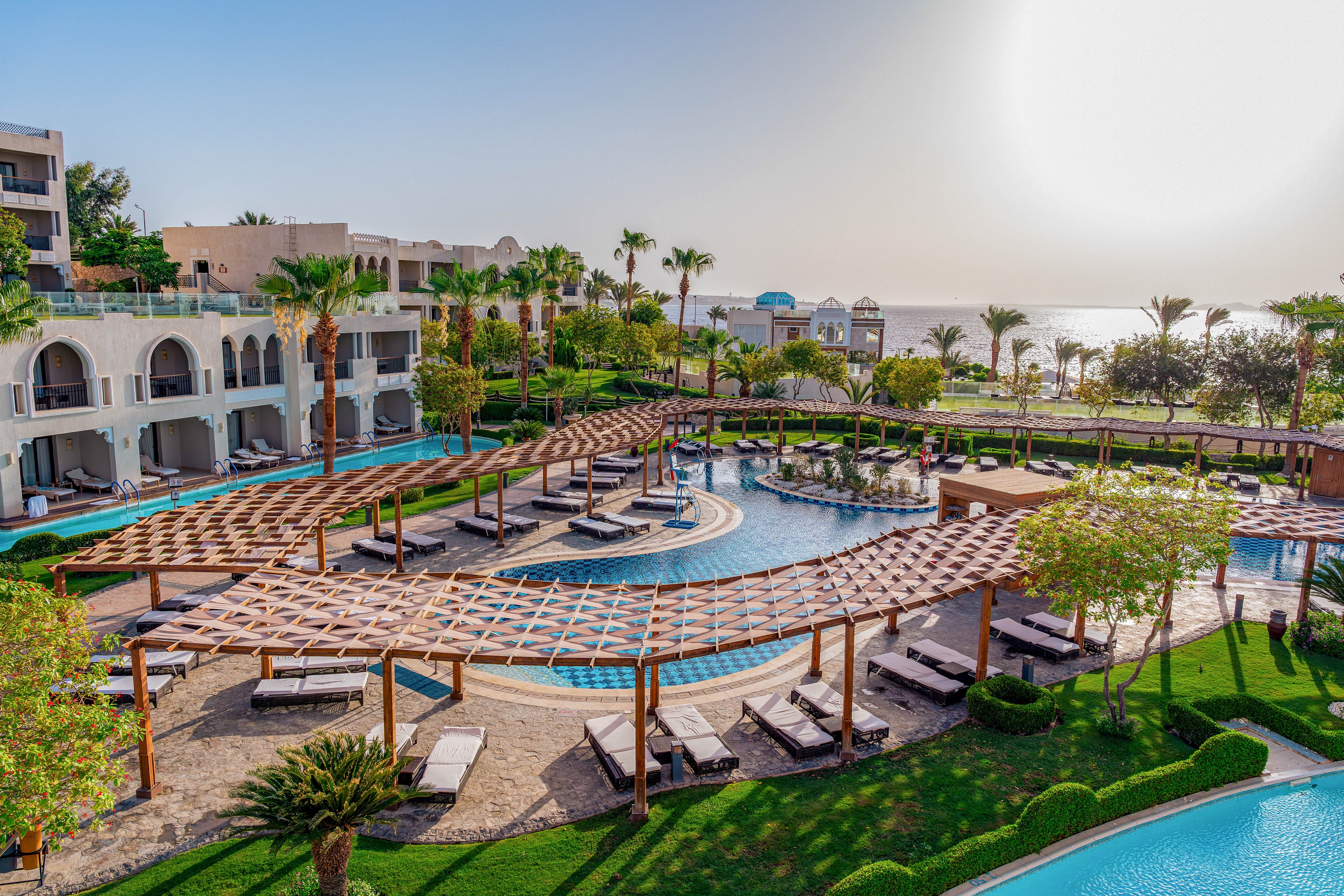 A luxurious seaside resort features a large, winding swimming pool surrounded by sun loungers and wooden pergolas. White multi-story buildings with balconies overlook the landscaped gardens and pool area. Palm trees and manicured lawns add a tropical feel to the scene. In the distance, the calm sea and bright rising sun create a serene vacation atmosphere.