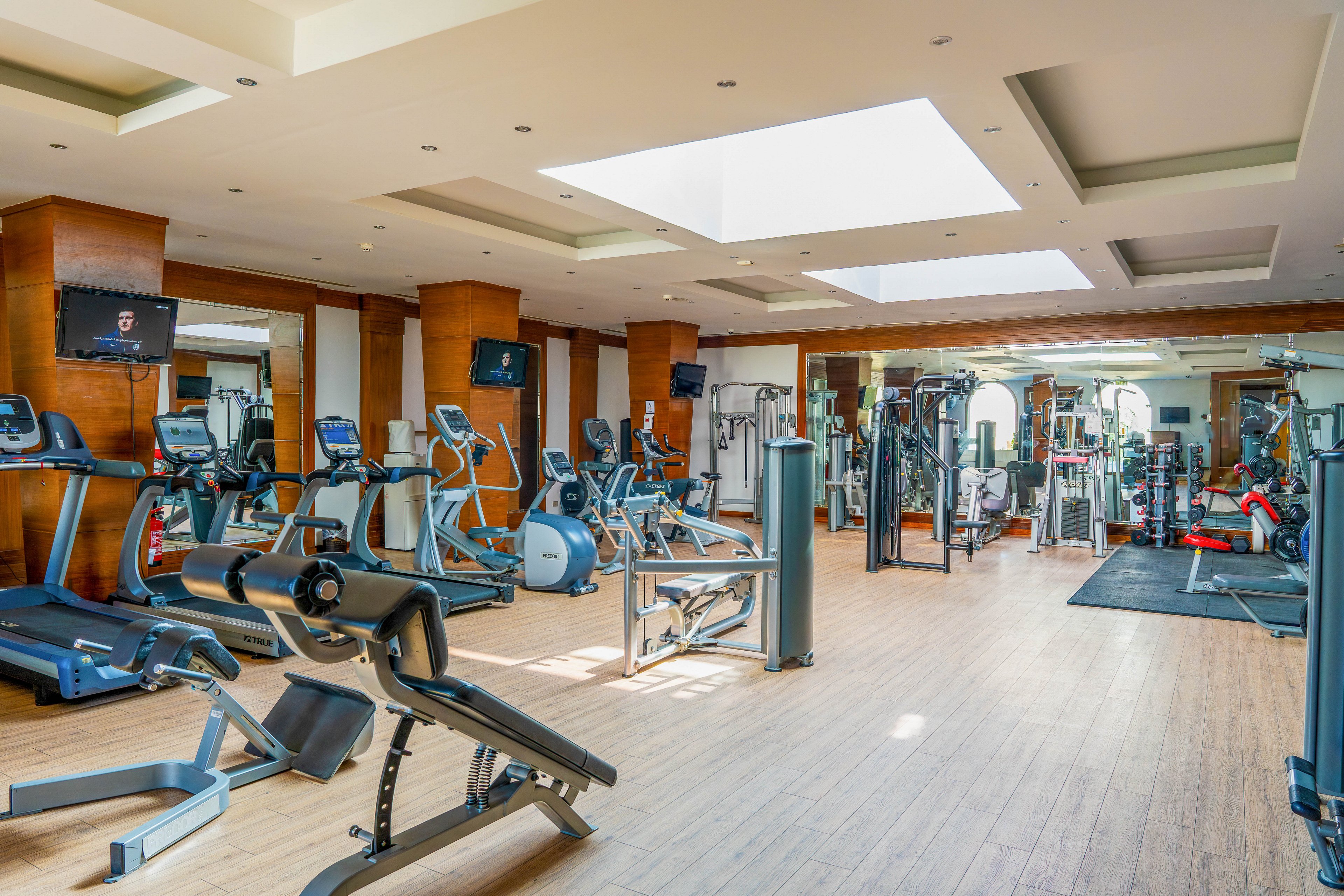 A spacious, modern gym interior features a variety of cardio and strength training machines neatly arranged. Treadmills, stationary bikes, and weight machines are positioned on a light wooden floor under large ceiling skylights. Wall-mounted televisions and mirrors enhance the bright, open atmosphere. The gym appears clean, organized, and ready for workouts, with no people present.