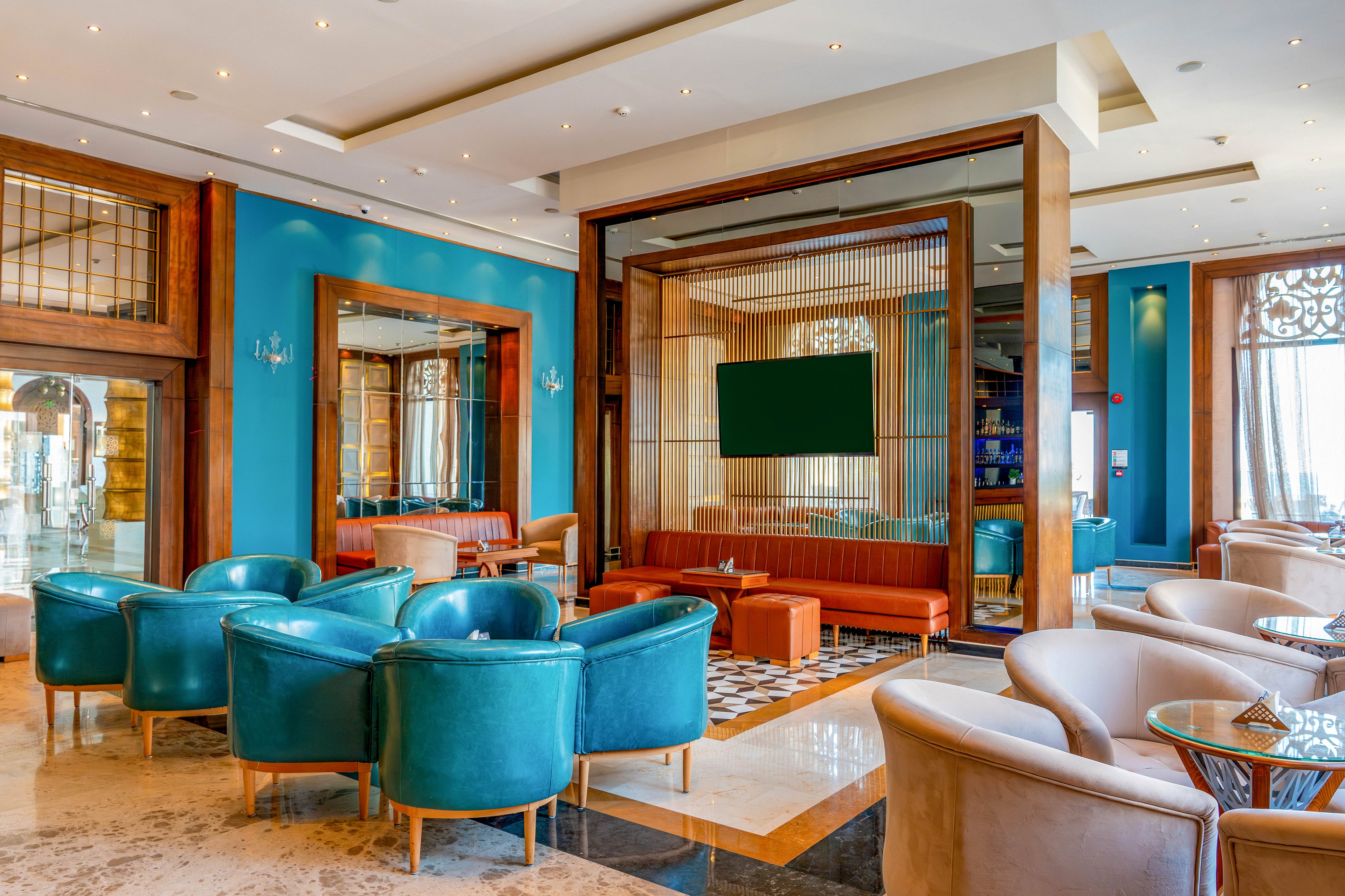 A contemporary hotel lounge features teal armchairs arranged around low tables on a polished floor. Warm wooden partitions frame a central seating area with orange leather sofas and a large blank screen on the wall. Turquoise walls, decorative metal grilles, and patterned rugs add visual interest to the space. Soft ceiling lighting and large windows create a bright, inviting atmosphere suitable for relaxation or informal meetings.