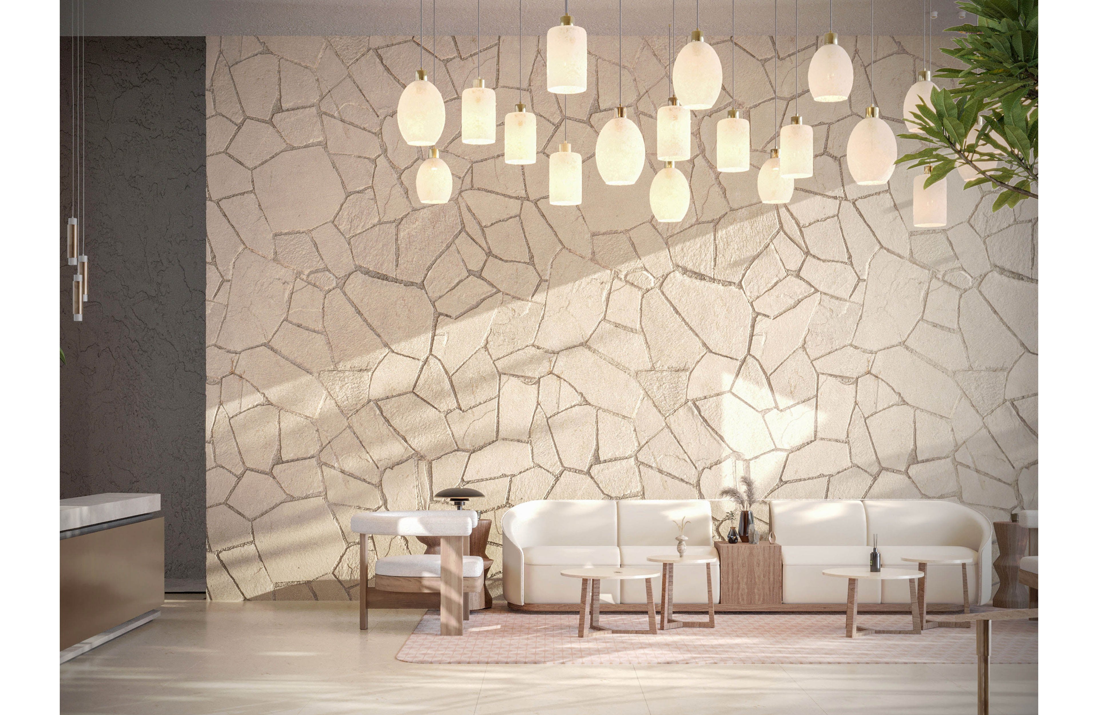 Contemporary lounge seating is arranged against a textured stone wall in a bright interior space. Multiple warm pendant lights hang from the ceiling, casting soft illumination across the room. Minimalist white sofas and small wooden tables create a clean, inviting atmosphere. A potted plant and subtle decor elements add a touch of nature to the modern design.