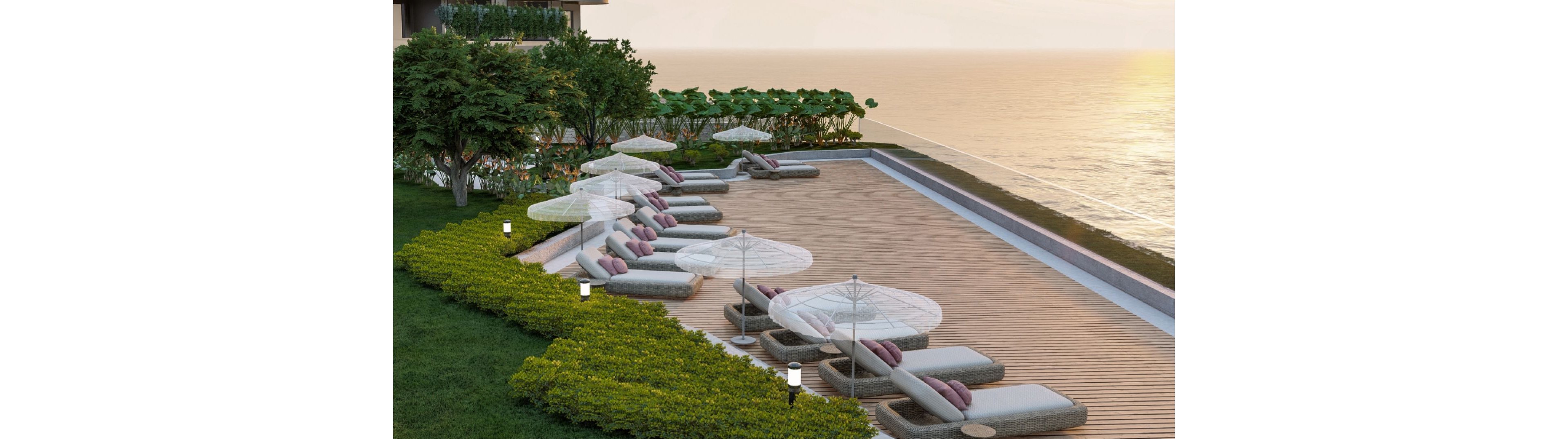 An elevated sun deck overlooks a calm ocean at sunset, featuring a row of cushioned loungers with white parasols. The seating area is bordered by neatly trimmed green shrubs and landscaped plants, creating a resort-like atmosphere. Soft, warm lighting from small pathway lamps enhances the tranquil mood. The scene suggests a peaceful, upscale coastal retreat designed for relaxation and leisure.