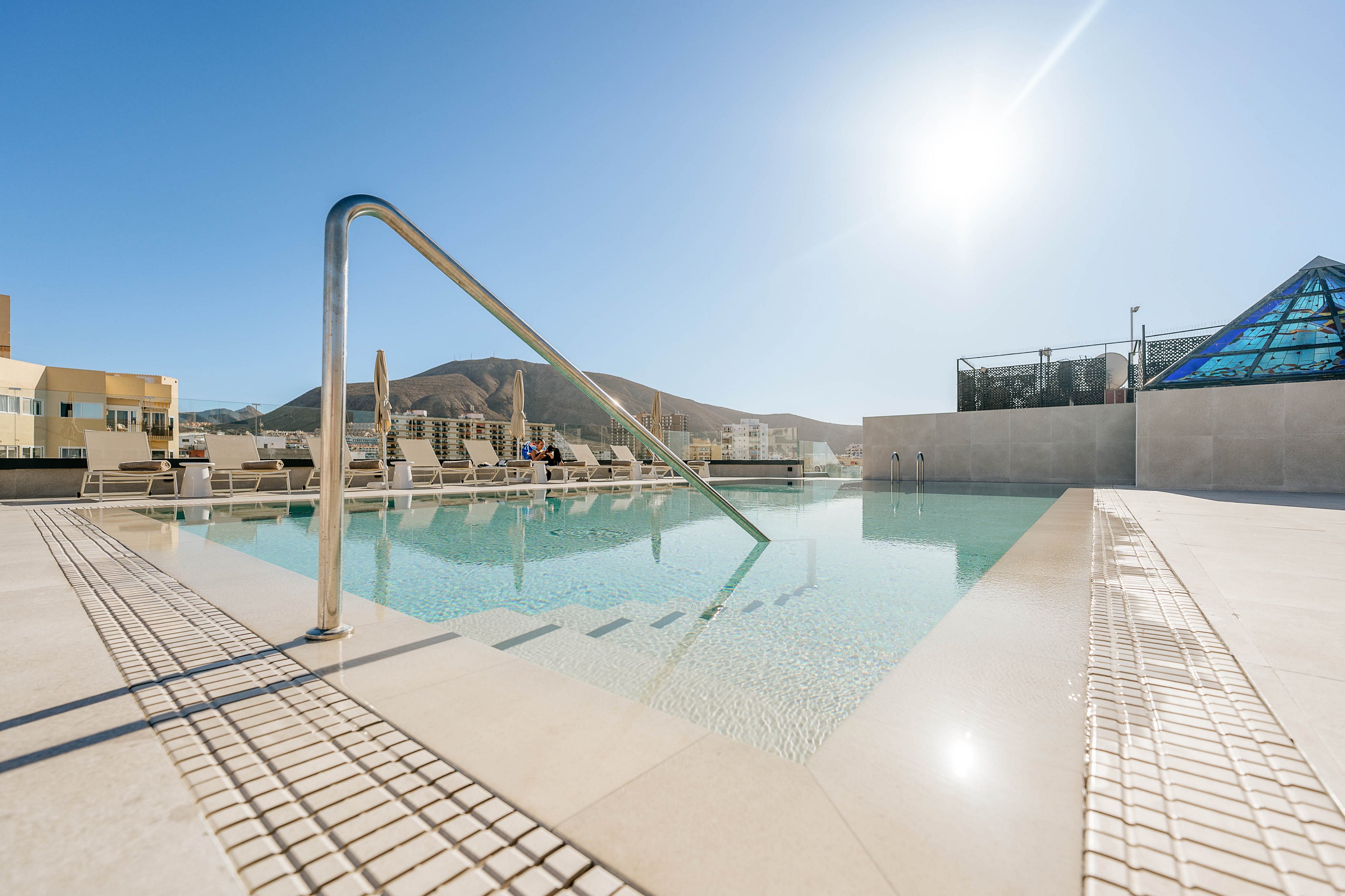 An outdoor rooftop swimming pool glistens under a bright, clear sky. Sun loungers and umbrellas line the pool deck, suggesting a relaxing leisure area. A metal handrail leads into the shallow water, with modern tiles and clean architectural lines surrounding the pool. Distant hills and contemporary buildings frame the background, emphasizing a resort or urban hotel setting.