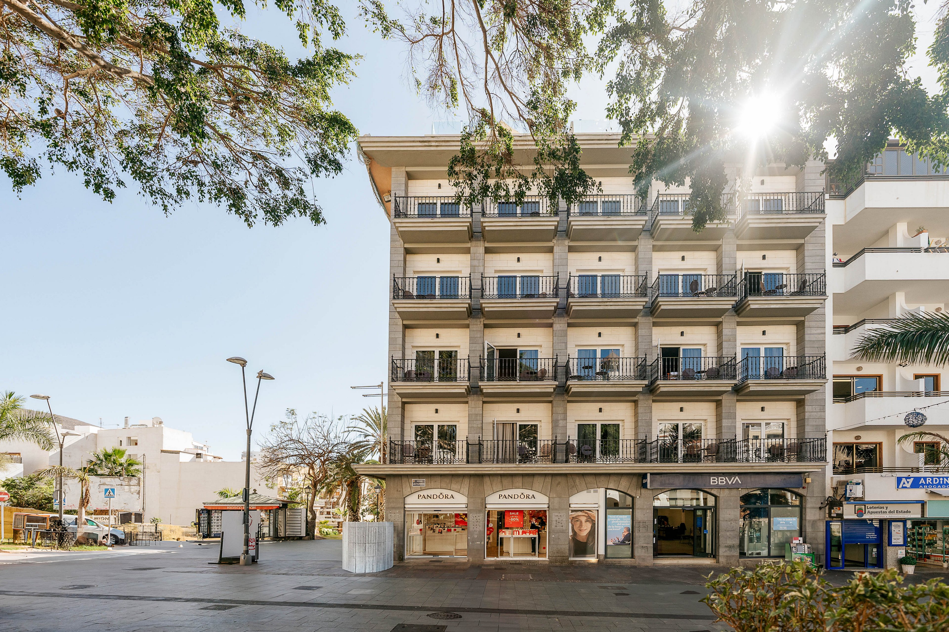 A mid-rise apartment building with multiple balconies stands along a sunny urban plaza. Ground-floor retail shops with large glass windows line the street level, while upper floors feature repeating balcony railings and doors. Trees frame the scene, and bright sunlight filters through the leaves, casting shadows on the open square. Nearby street furniture and adjacent buildings suggest a lively city neighborhood.