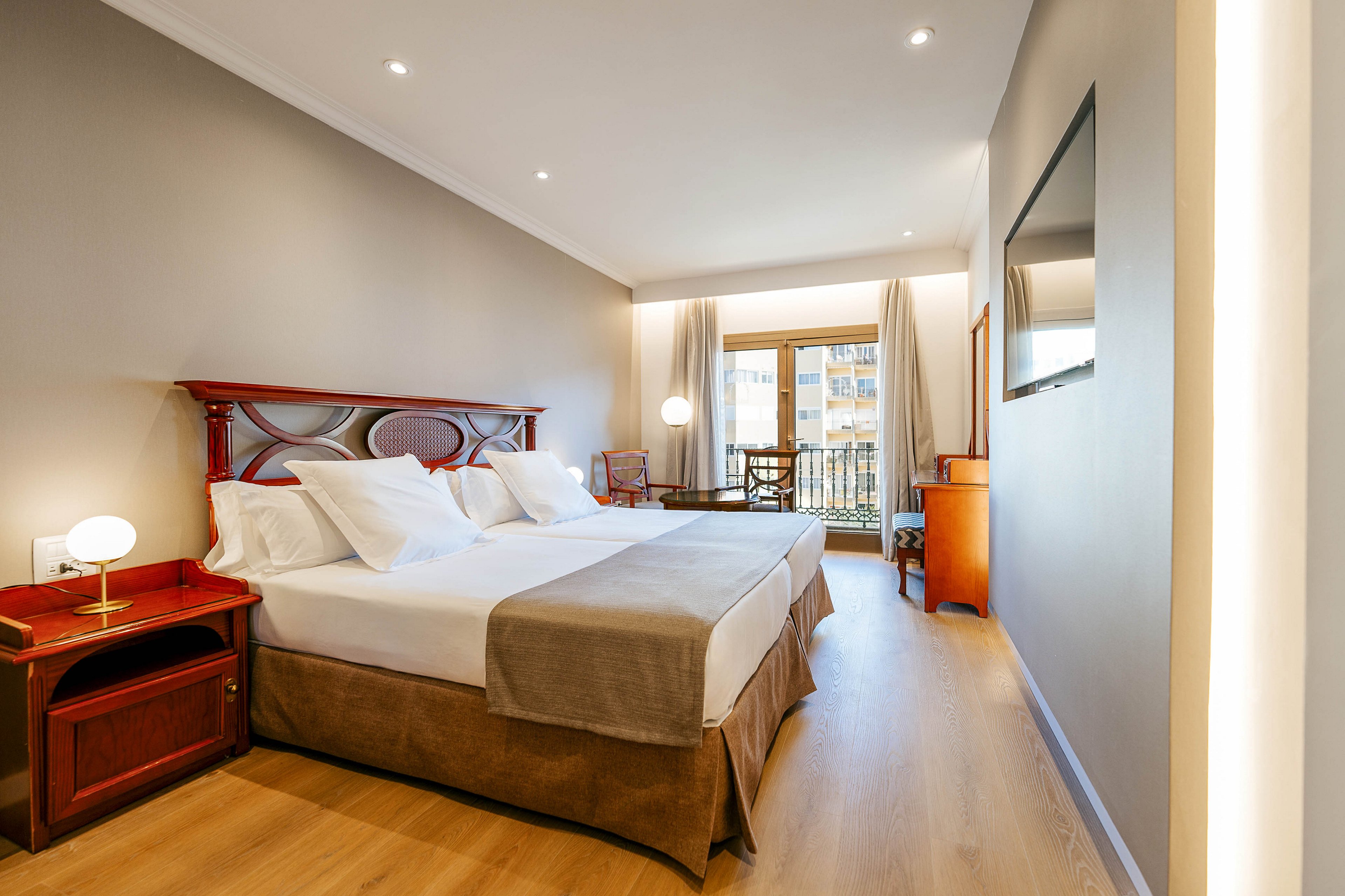 A contemporary hotel room features two neatly made twin beds with white linens and beige throws. Warm wooden furniture, including bedside tables and a desk, adds contrast to the neutral walls and light wood flooring. Recessed ceiling lights and a small bedside lamp create a bright yet cozy atmosphere. A large window with curtains opens to a balcony, bringing in natural light and an exterior city view.