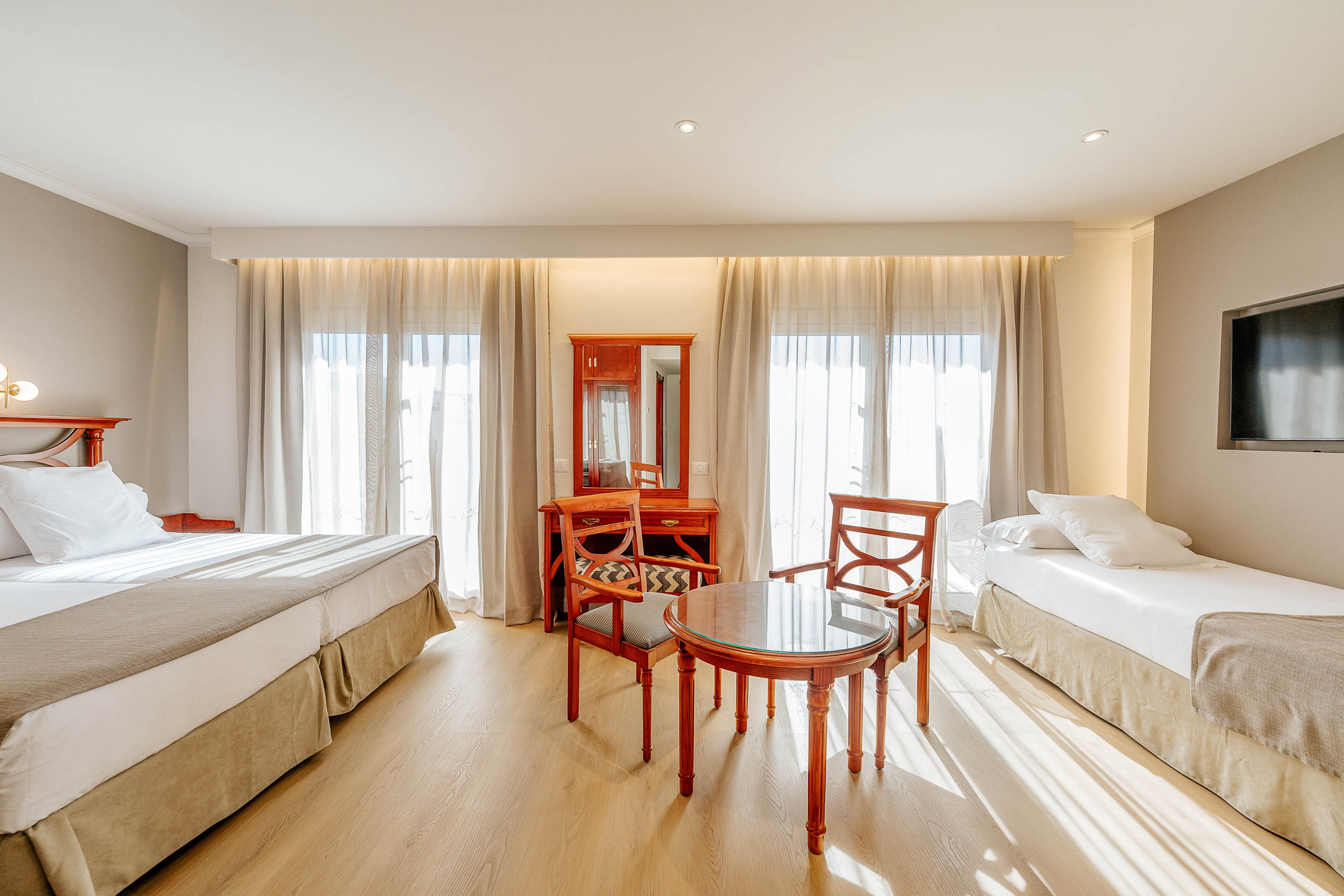 Spacious hotel room featuring a large double bed and a single bed with white linens. A round wooden table with chairs sits in the center of the room near a wooden vanity and mirror. Floor-to-ceiling windows with sheer curtains allow abundant natural light to fill the space. Neutral walls and light wood flooring create a clean, minimalist atmosphere, complemented by a wall-mounted flat-screen television.