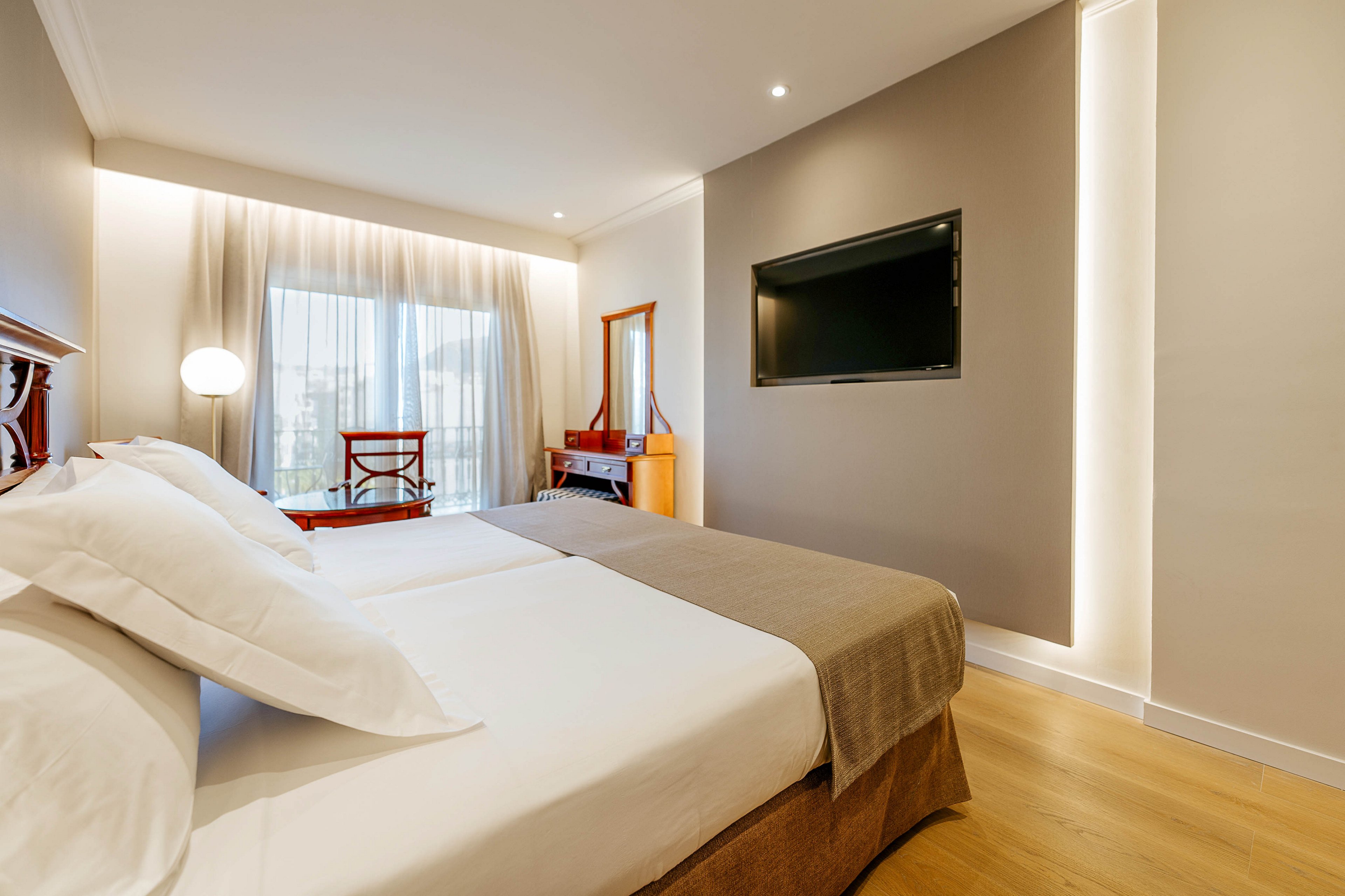 A bright, modern hotel room features two neatly made twin beds with white linens and beige throws. A large flat-screen television is mounted on a taupe accent wall with integrated strip lighting. Natural light enters through a large window covered with sheer curtains, illuminating a wooden desk and chair in the background. The space has a clean, minimalist design with warm wood flooring and recessed ceiling lights.
