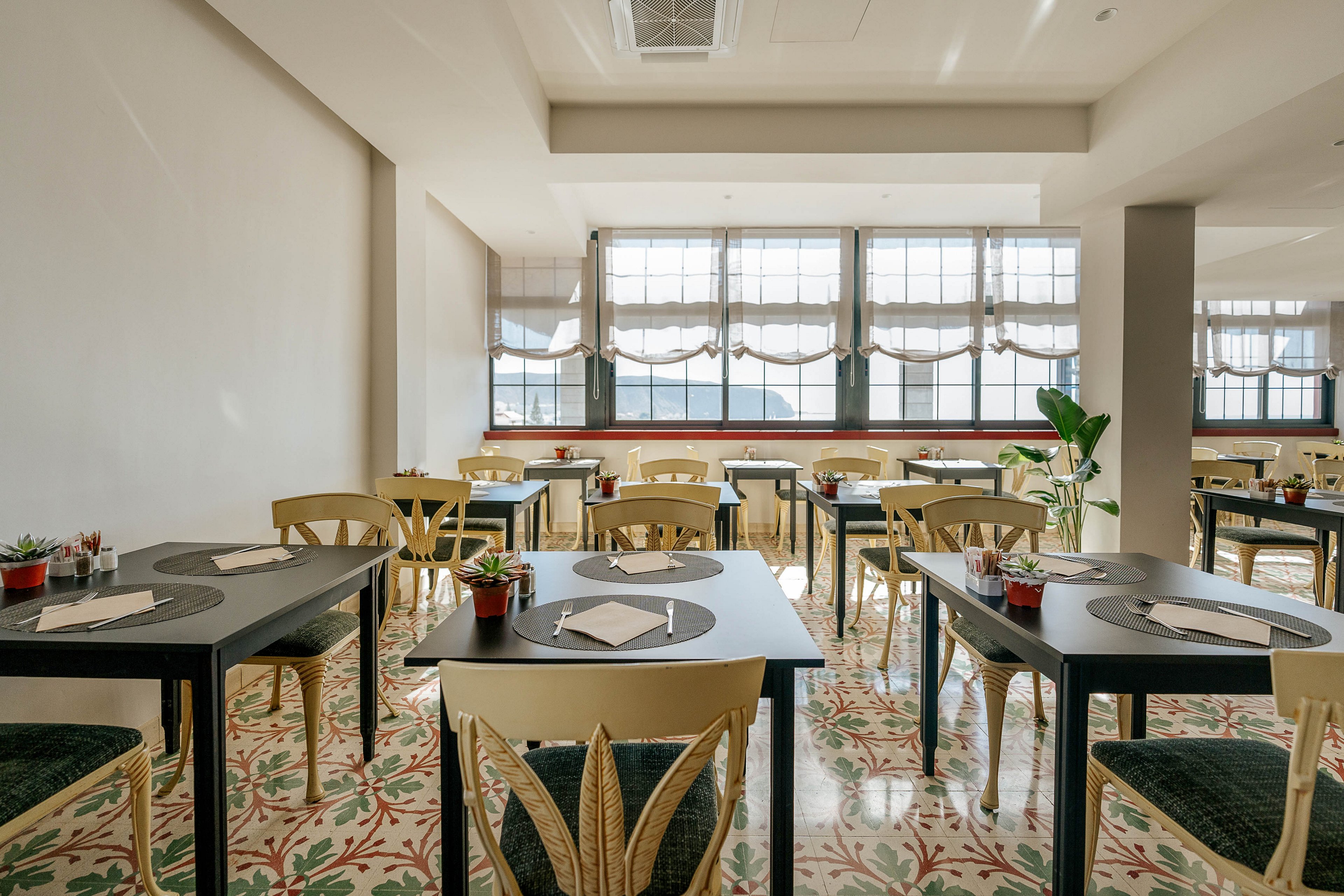 A bright, modern restaurant dining room is shown with neatly arranged tables and chairs. Each dark table has placemats, folded napkins, and small potted plants as simple decor. Large windows with sheer curtains let in abundant natural light, illuminating the patterned tile floor. The space appears clean, quiet, and ready to welcome guests.