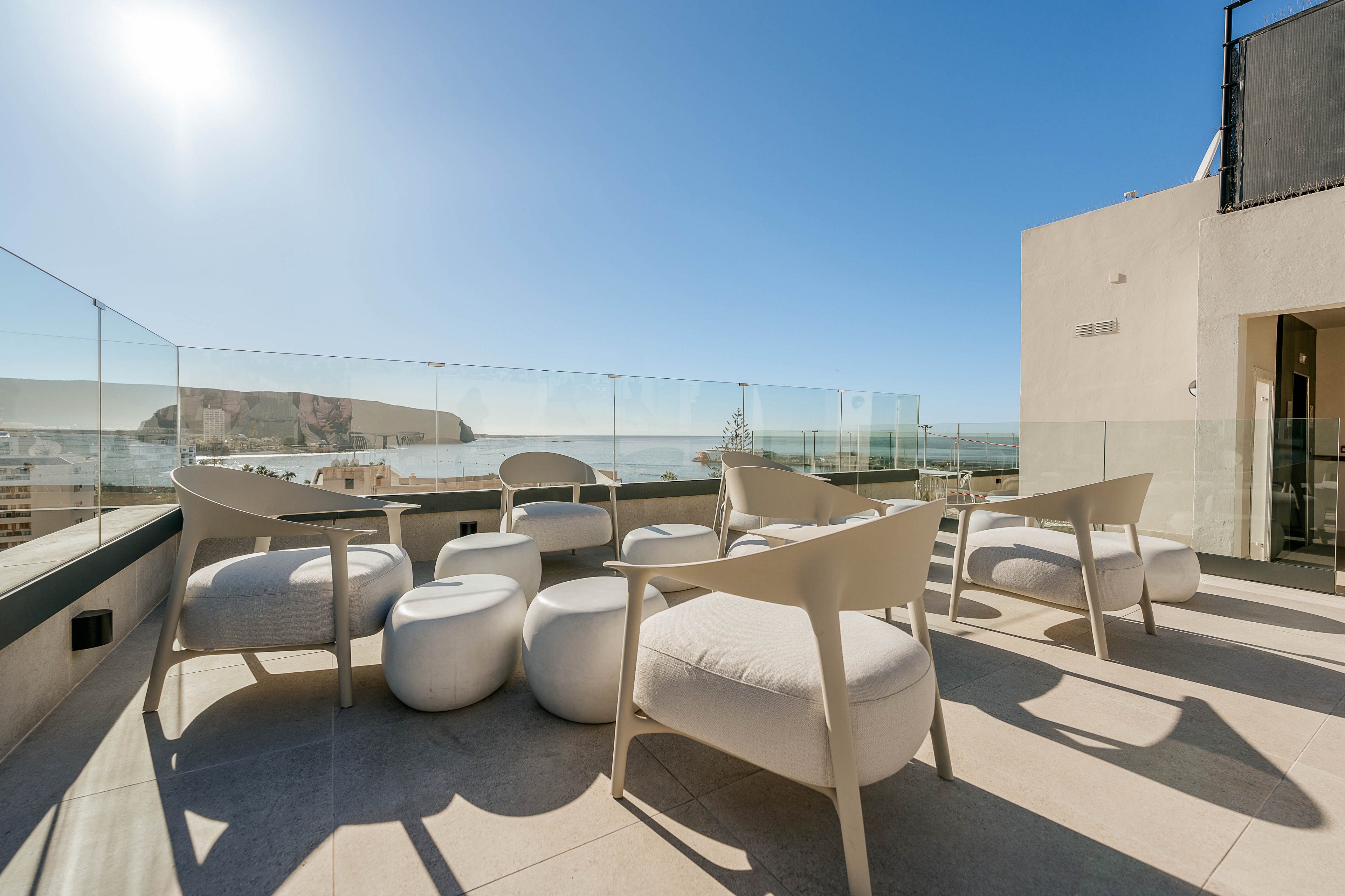 A contemporary rooftop terrace features minimalist chairs and cushioned stools arranged for outdoor lounging. Glass railings surround the space, opening to a clear view of the sea and distant coastline. Bright sunlight and a cloudless blue sky create a warm, inviting atmosphere. The clean architectural lines and neutral tones emphasize a modern, luxurious setting.