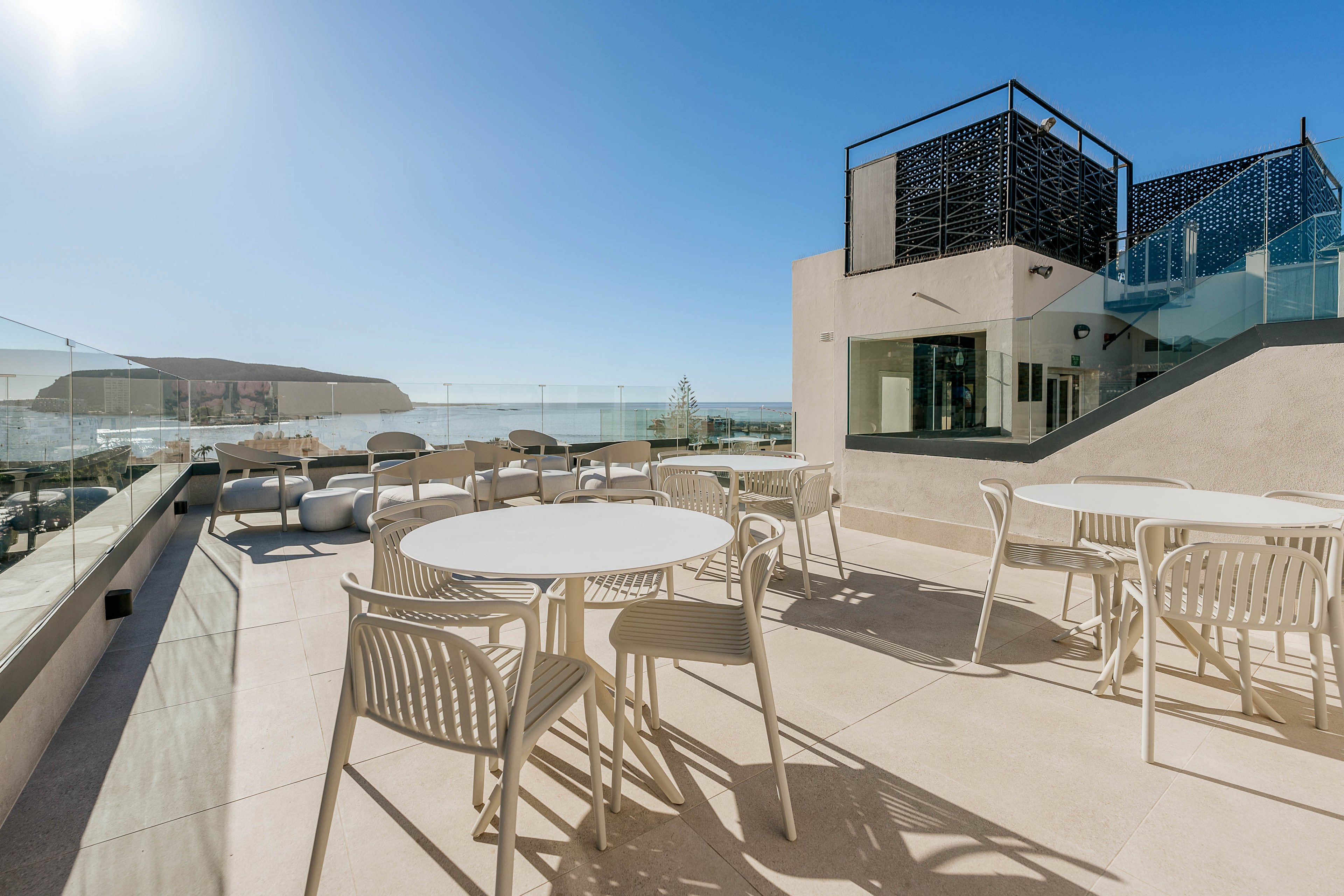 A spacious rooftop terrace features several round white tables and contemporary chairs under a clear blue sky. Glass railings line the edges, offering an unobstructed view of the ocean and distant hills. A modern building structure with geometric design elements rises on one side of the terrace. The bright sunlight and clean architectural lines create an airy, minimalist atmosphere ideal for relaxation or social gatherings.