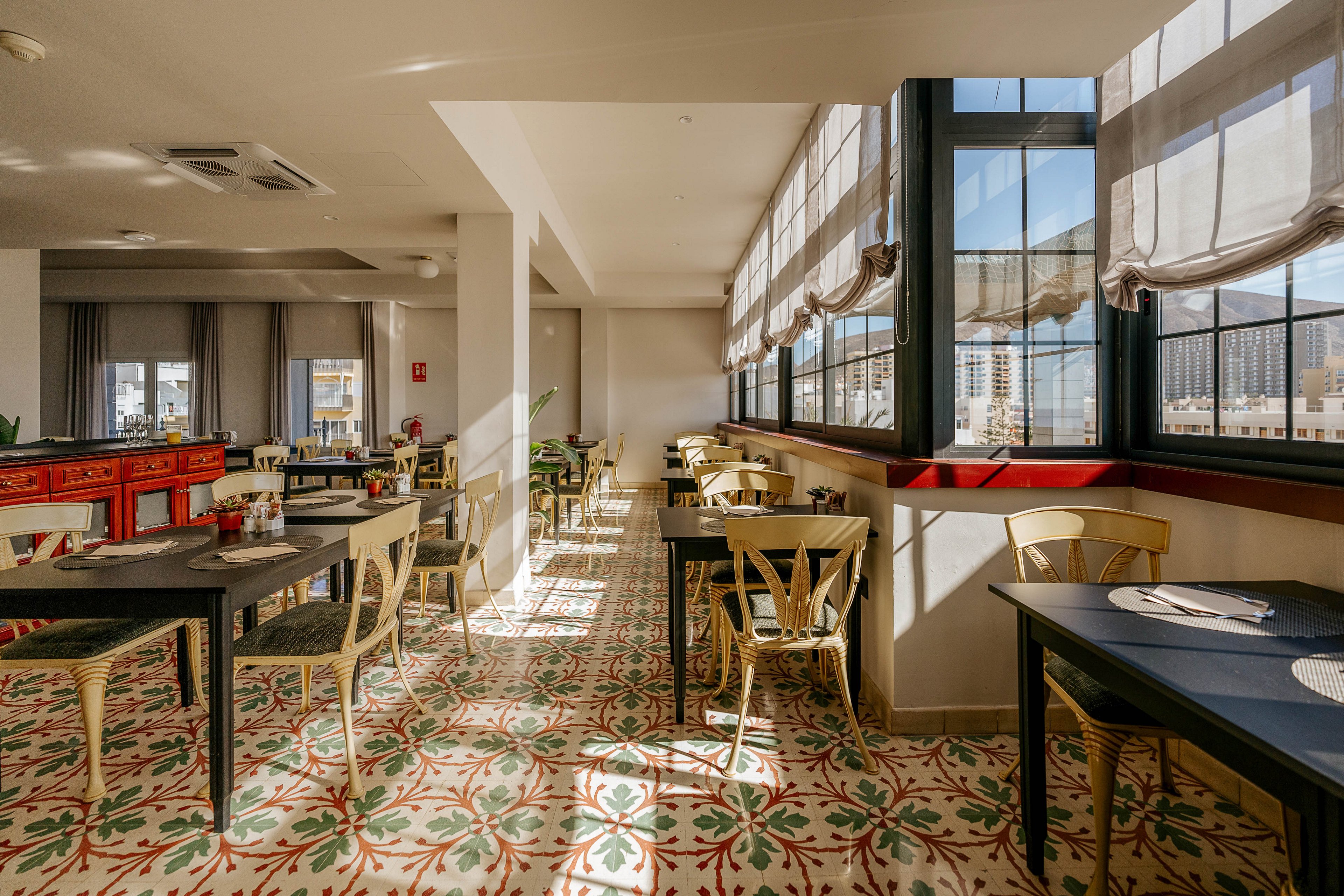 A bright restaurant dining room is filled with natural light streaming through large windows. Empty tables and chairs are neatly arranged, with place settings ready for guests. The patterned tile floor adds a decorative touch with colorful geometric designs. Soft curtains and neutral walls create a warm, inviting atmosphere in the space.