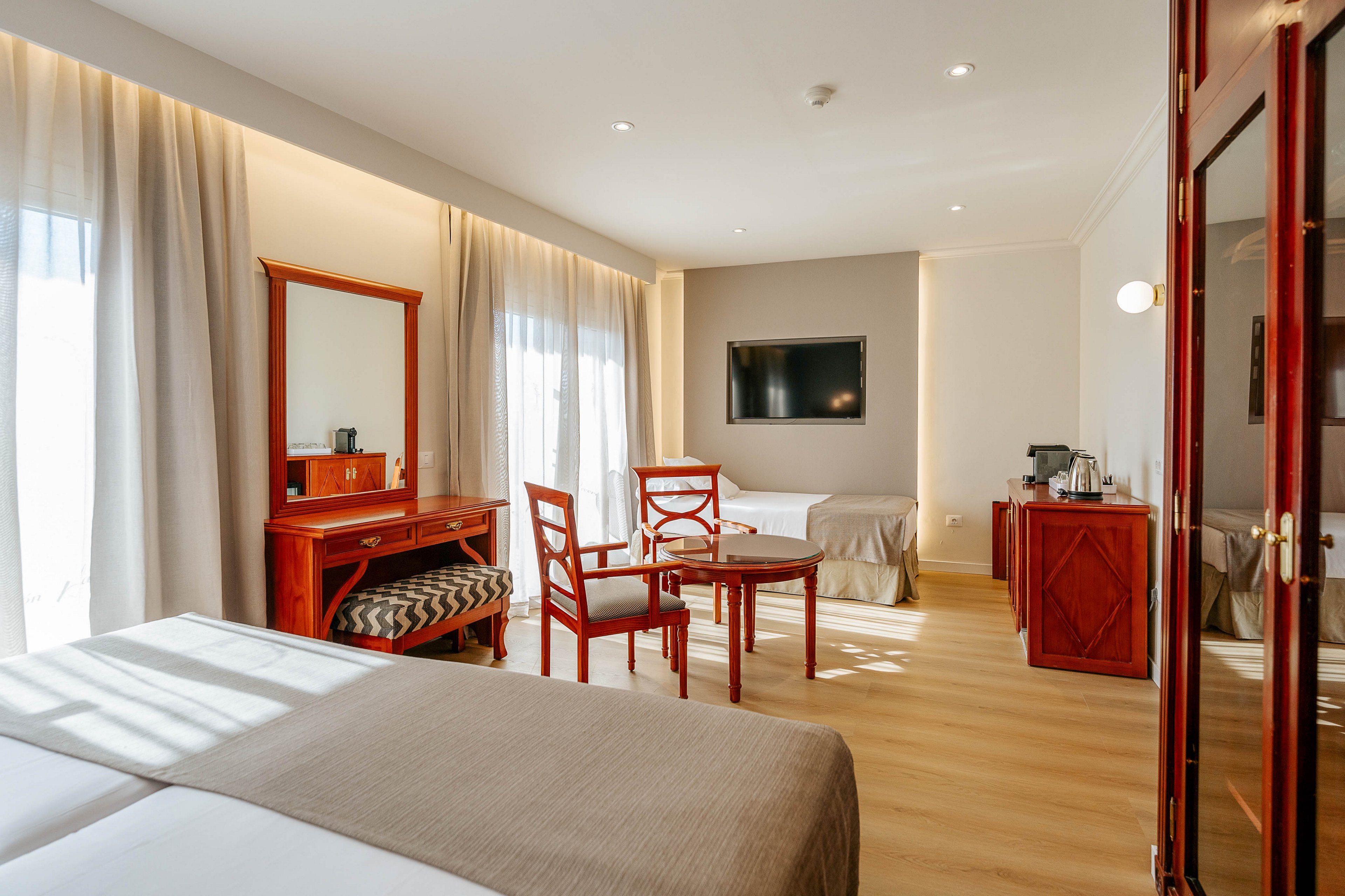 A spacious hotel room features two neatly made beds in the foreground. Warm wooden furniture, including a table with chairs and a vanity desk with a cushioned bench, adds a classic touch. Large windows with sheer curtains fill the space with natural light, complementing the neutral walls and light wood flooring. A flat-screen TV is mounted on the wall opposite the beds, and a cabinet with a coffee station sits in the corner, creating a comfortable guest environment.