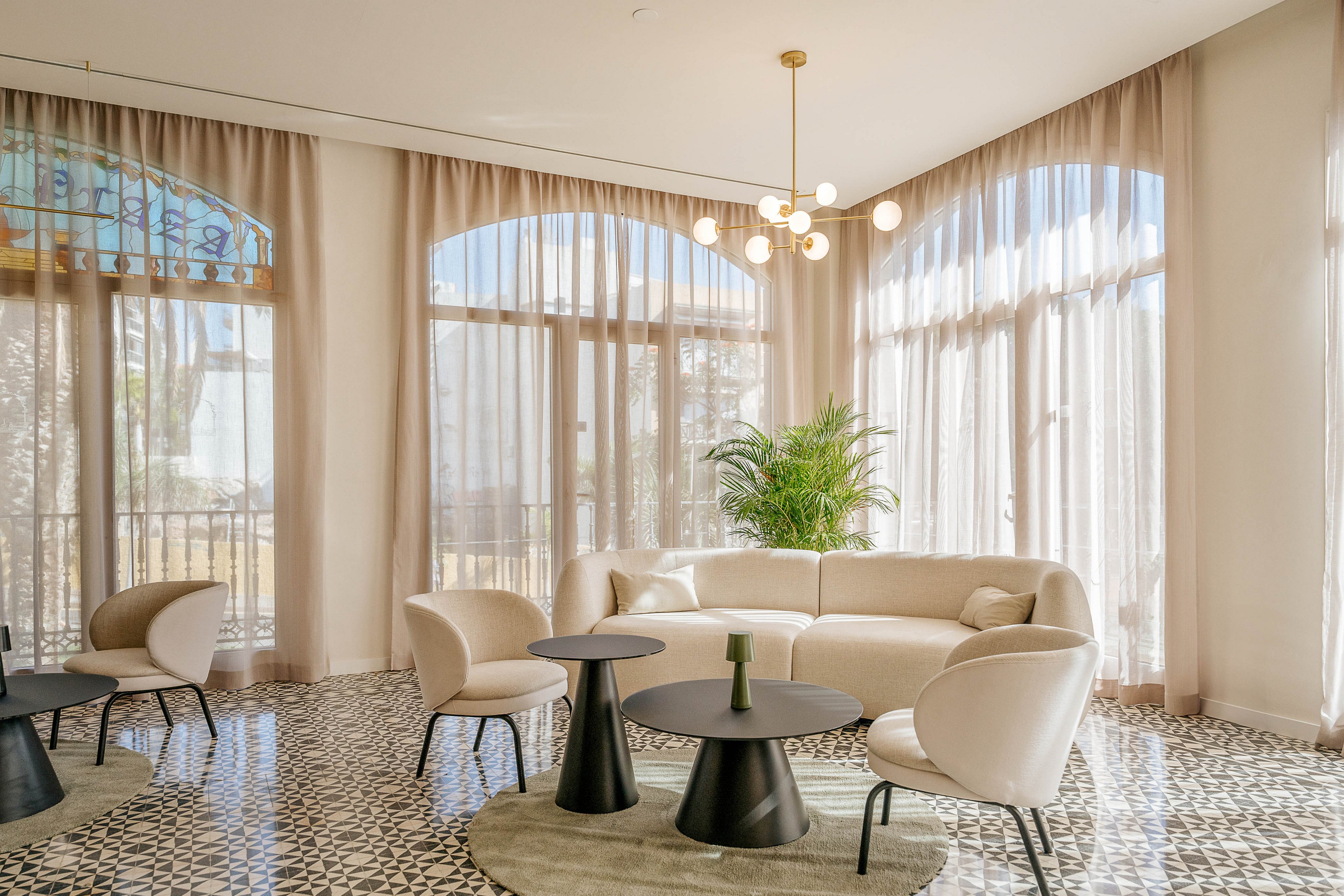 A contemporary lounge area features a cream-colored sofa and matching armchairs arranged around small round tables. Large arched windows with sheer curtains fill the room with natural light. A modern pendant light with multiple globes hangs from the ceiling above the seating area. Patterned floor tiles and a tall green plant add texture and contrast to the otherwise neutral interior.