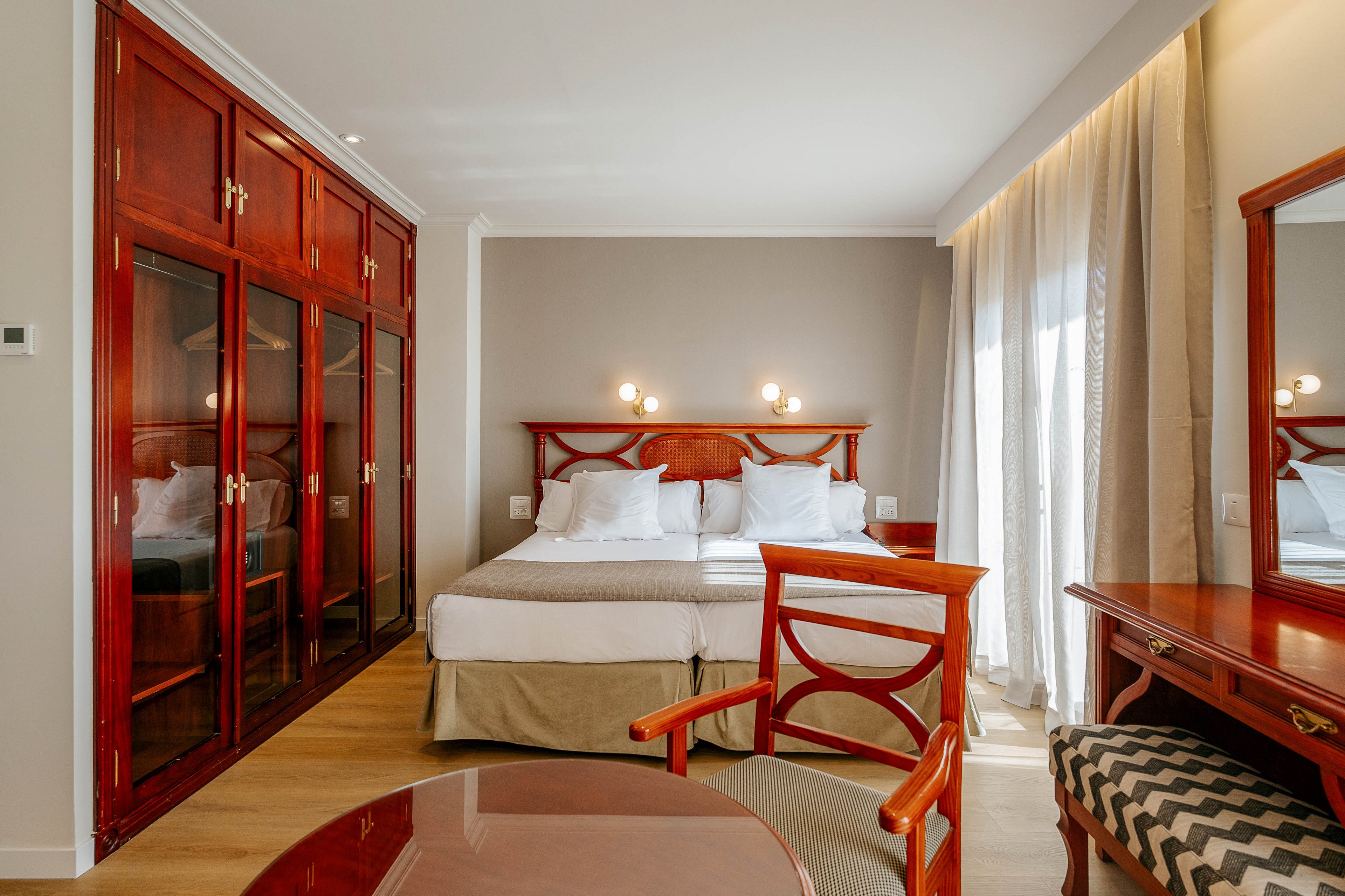 A neatly made double bed with white linens stands against a neutral wall in a bright hotel room. Classic dark wood furniture, including a wardrobe with glass doors, a desk, and chairs, frames the space. Large windows with sheer curtains allow natural light to fill the room. The overall atmosphere is clean, orderly, and inviting, suitable for comfortable accommodation.
