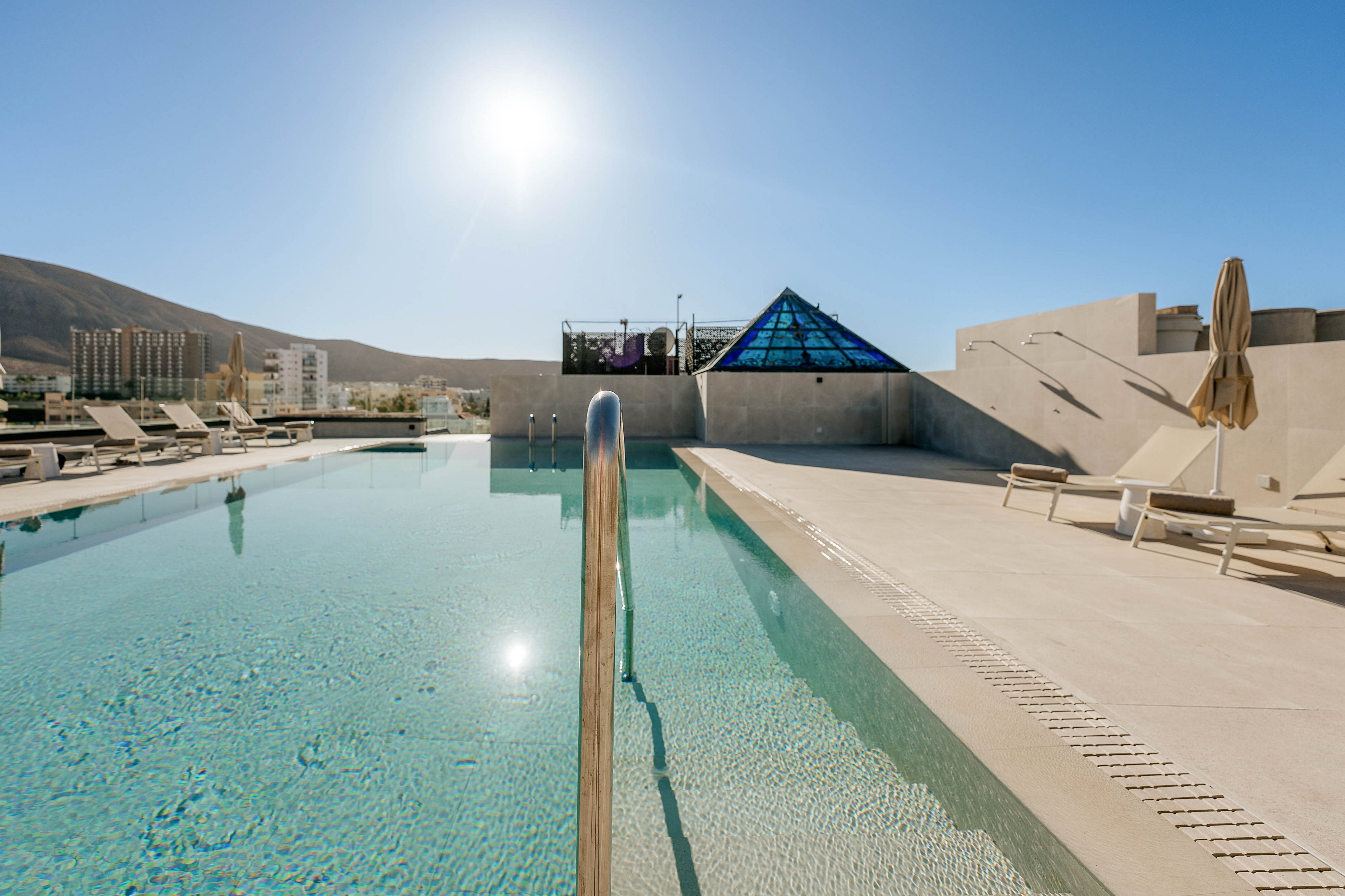 A clear rooftop swimming pool stretches into the distance under a bright midday sun. Lounge chairs and umbrellas line the pool deck, suggesting a relaxing resort or hotel setting. In the background, a glass pyramid structure and low modern buildings frame the scene, with hills visible beyond. The calm water and minimalistic architecture create a serene, luxurious atmosphere.
