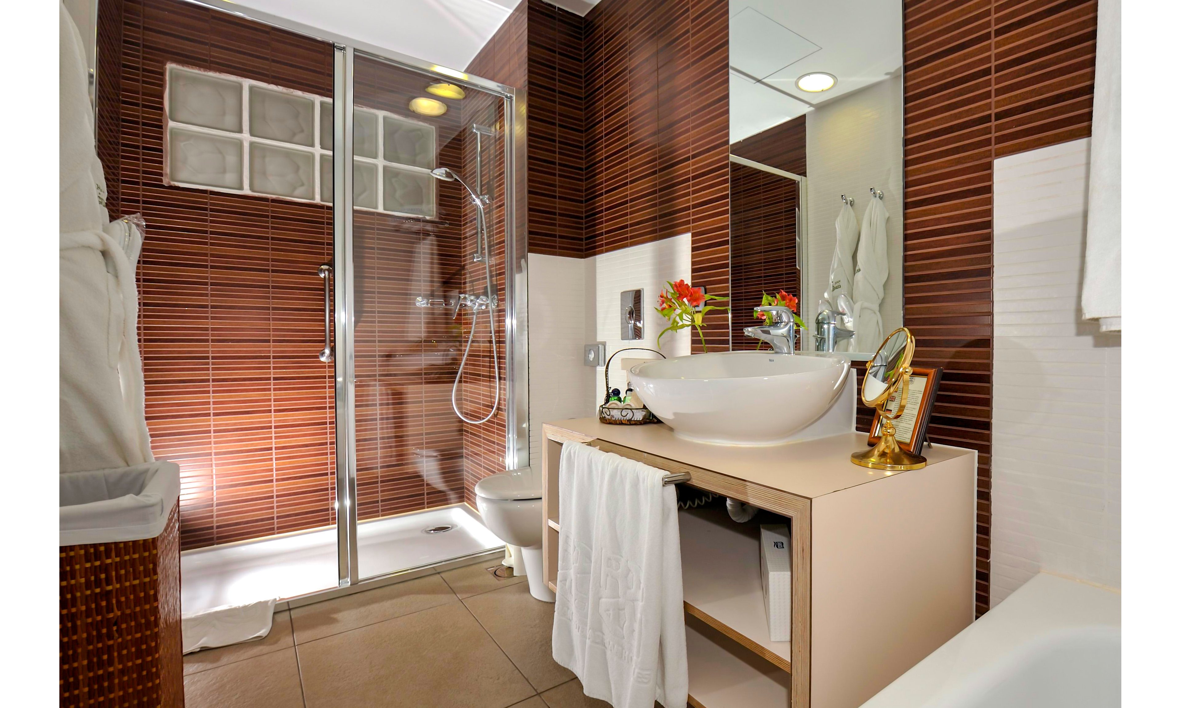 A contemporary bathroom featuring a glass shower enclosure and a sleek white sink on a wooden vanity. The walls are adorned with horizontal wooden paneling, creating a warm and inviting atmosphere. Fresh flowers and a decorative mirror add a touch of elegance to the space.