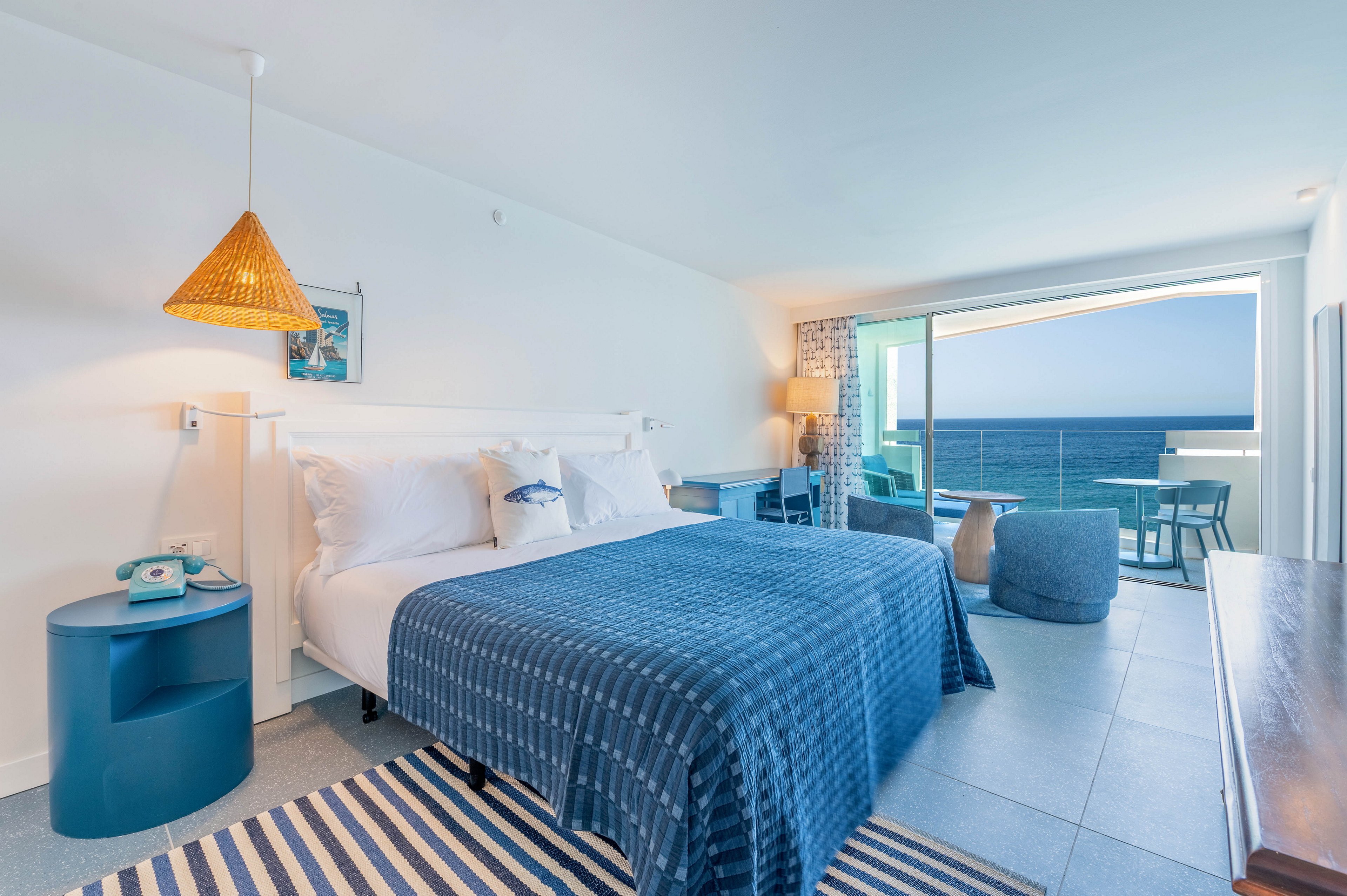 A modern hotel bedroom features a large bed with blue patterned bedding and white pillows. The room is decorated in a coastal style with blue furniture, a striped rug, and a woven pendant lamp. A sliding glass door opens to a balcony overlooking the ocean, bringing in abundant natural light. The overall atmosphere is airy, relaxed, and inviting for seaside vacation stays.