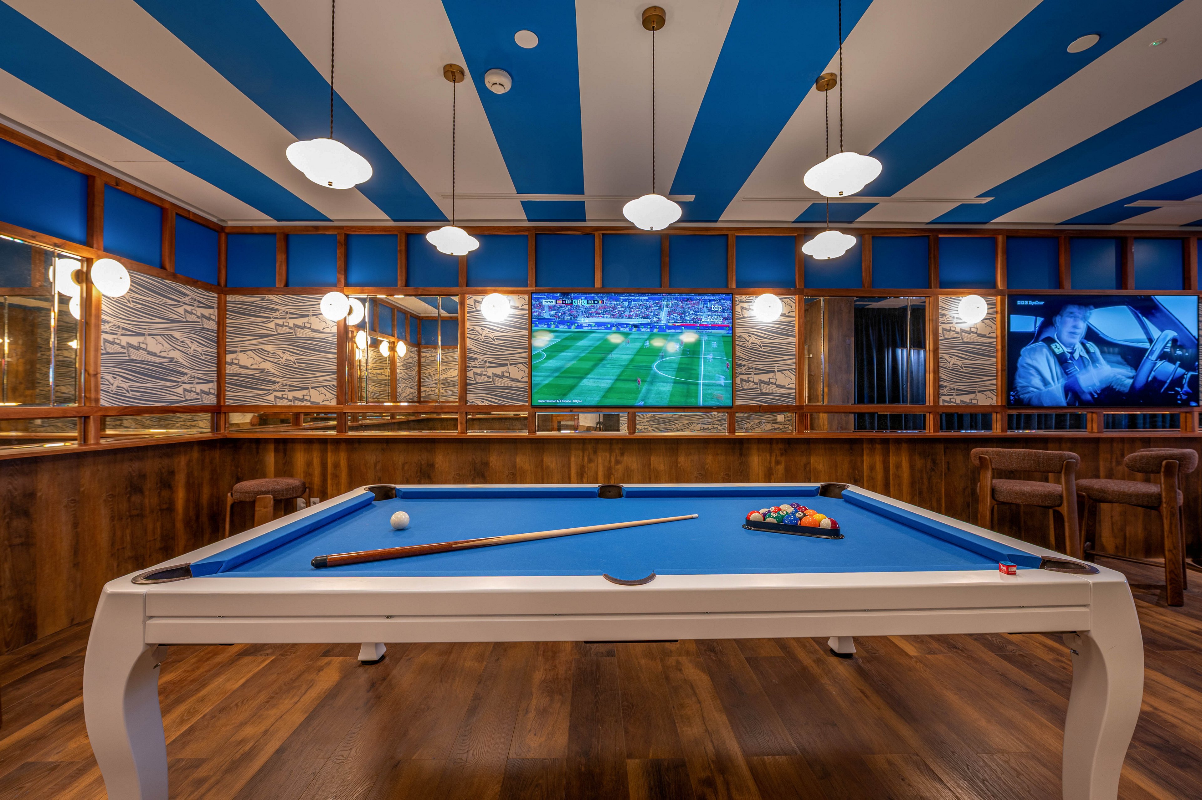 A contemporary game room features a white and blue pool table centered on a wooden floor. A cue stick and racked billiard balls rest on the bright blue felt surface. The room is surrounded by wood paneling, patterned wall sections, and large screens displaying a soccer match and a monochrome scene. Overhead pendant lights and a striped blue and white ceiling create a stylish, inviting atmosphere.