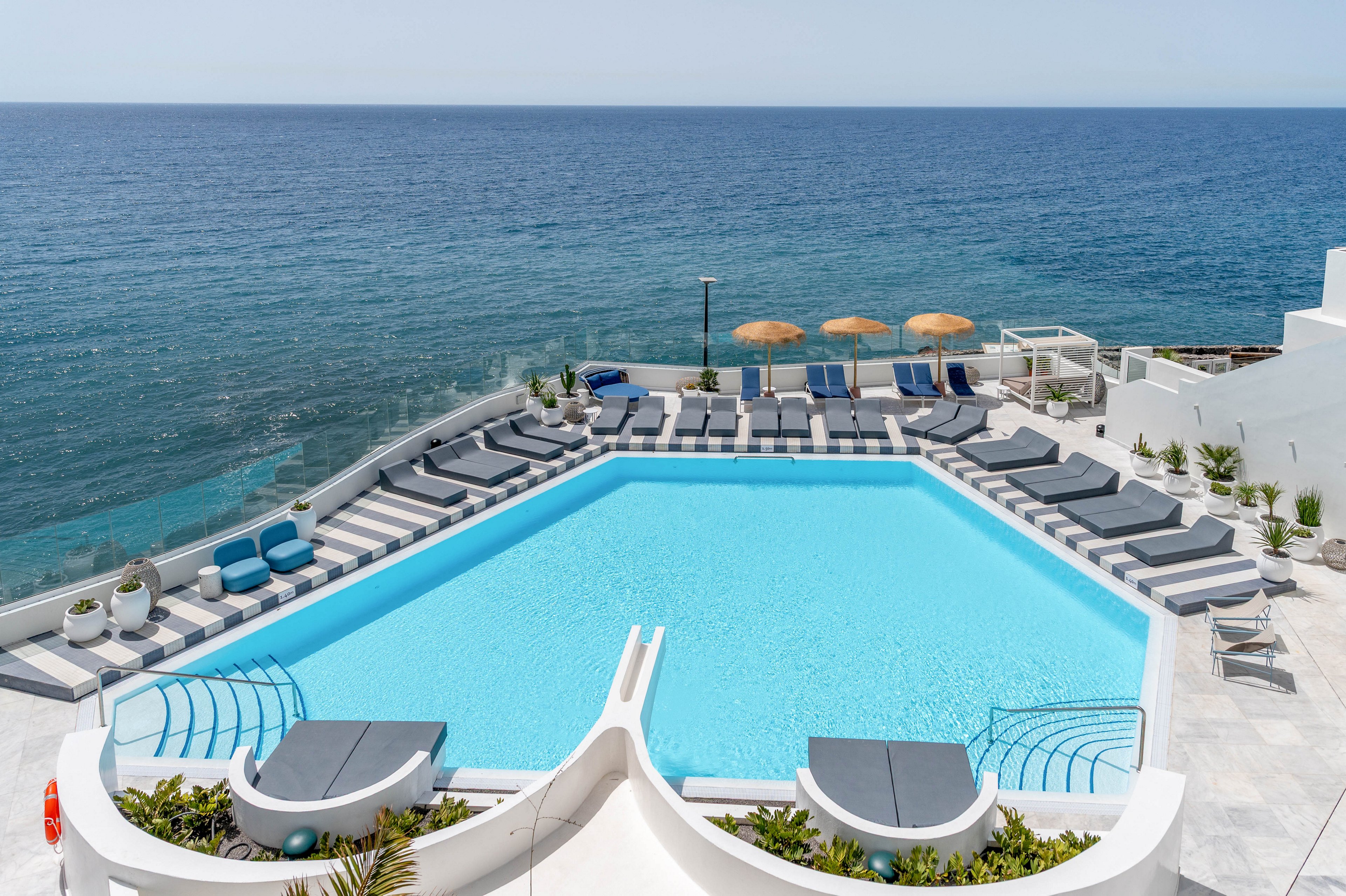 A bright blue swimming pool sits on a white terrace overlooking a calm ocean. Numerous gray sun loungers and umbrellas surround the pool, arranged neatly along the edges. The setting appears to be a modern coastal resort with minimalist white architecture and clean lines. The overall atmosphere is tranquil and inviting, ideal for sunbathing and relaxing by the sea.