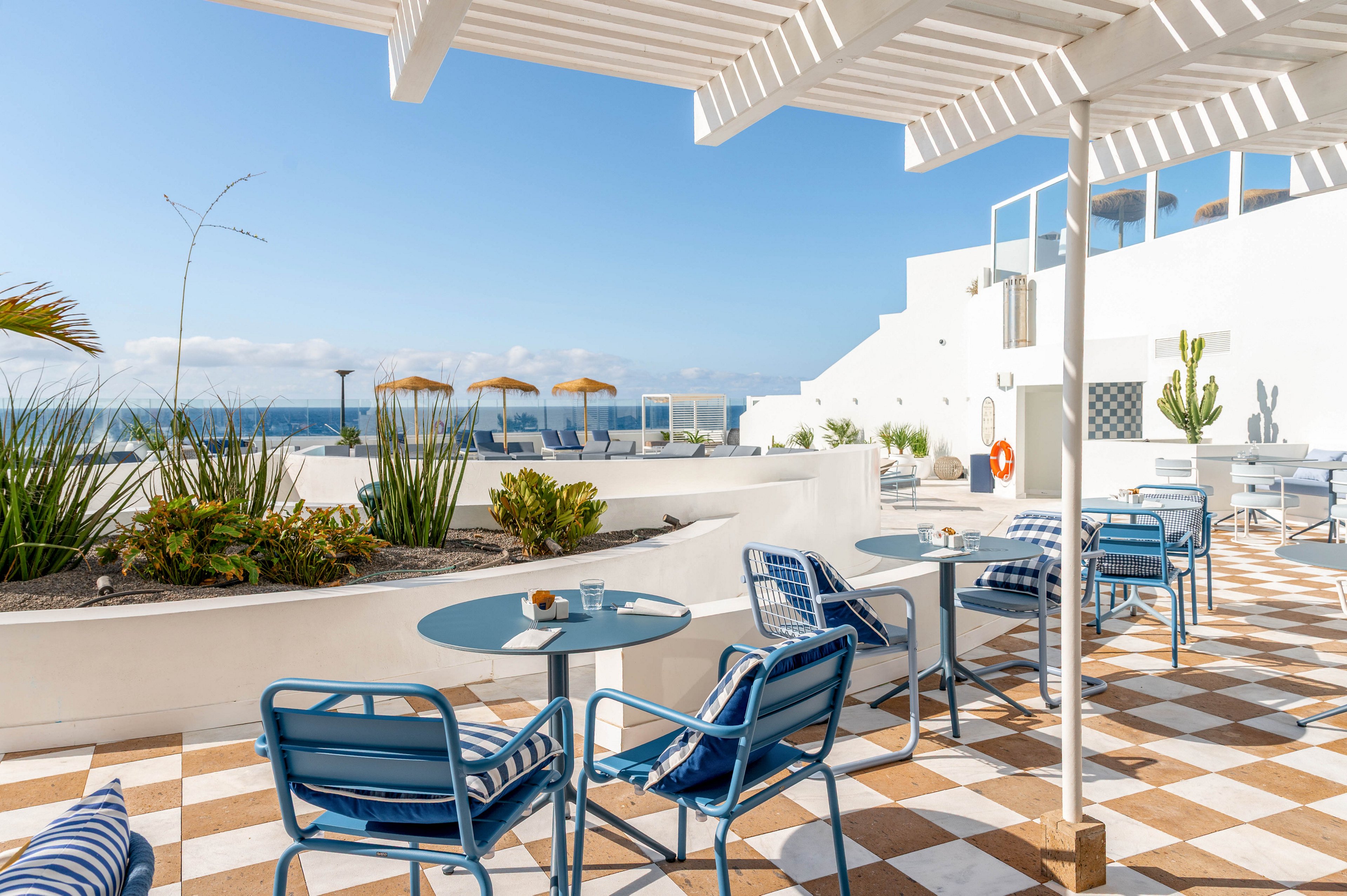 An outdoor terrace features blue metal lounge chairs and round glass tables under white pergolas. The space overlooks the ocean with straw parasols visible in the distance and a clear blue sky above. Checkerboard tiles in beige and white cover the floor, complemented by white walls and built-in planters with green plants and cacti. Cushions and striped textiles on the chairs add a relaxed, coastal atmosphere to the bright, minimalist setting.
