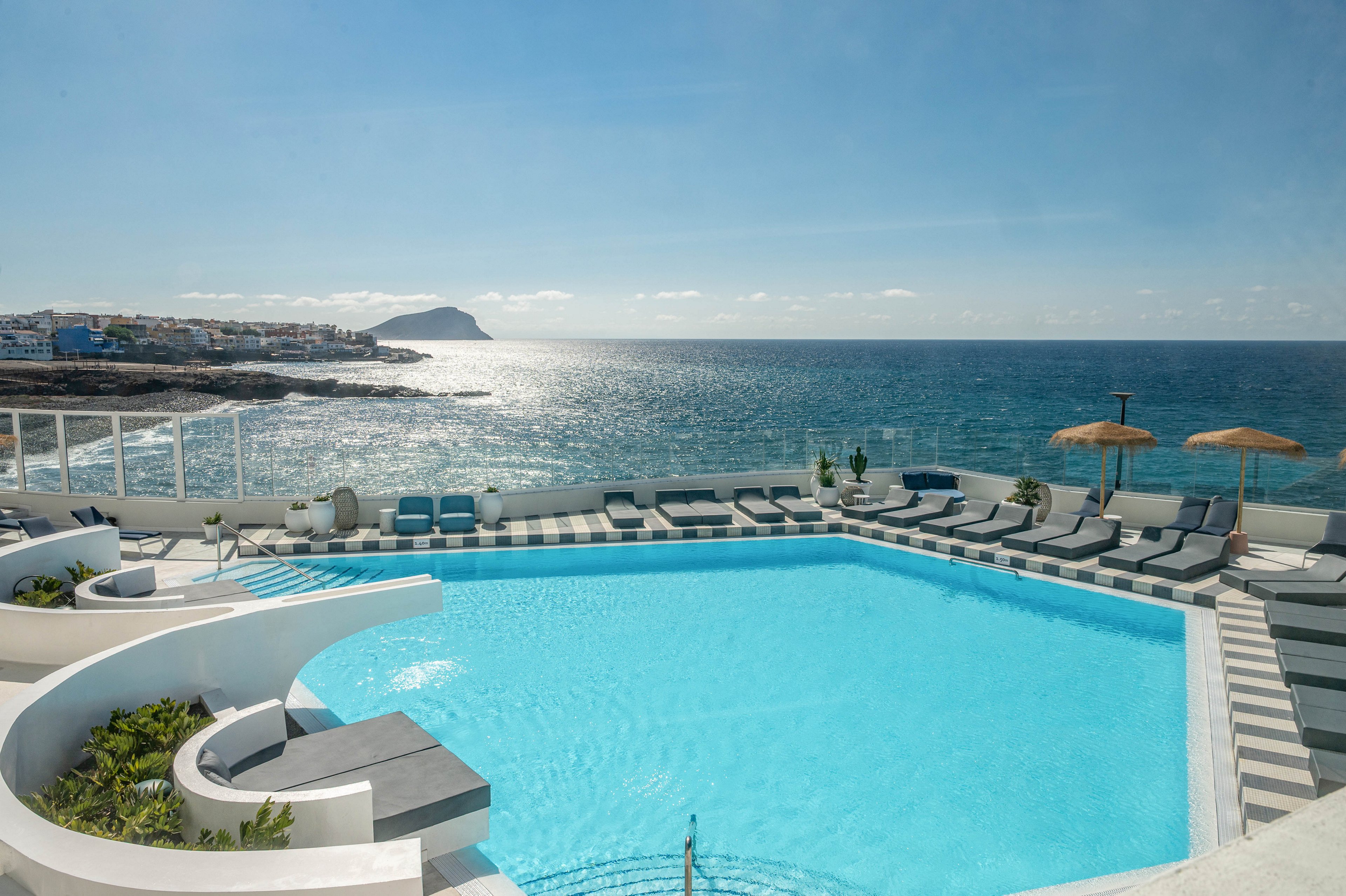A contemporary outdoor swimming pool sits on a terrace directly above the sea. Sun loungers and umbrellas line the curved deck, facing the sparkling water and distant coastline. The calm ocean horizon and clear sky create a serene vacation atmosphere. The scene suggests a luxury resort setting ideal for relaxation and sunbathing.