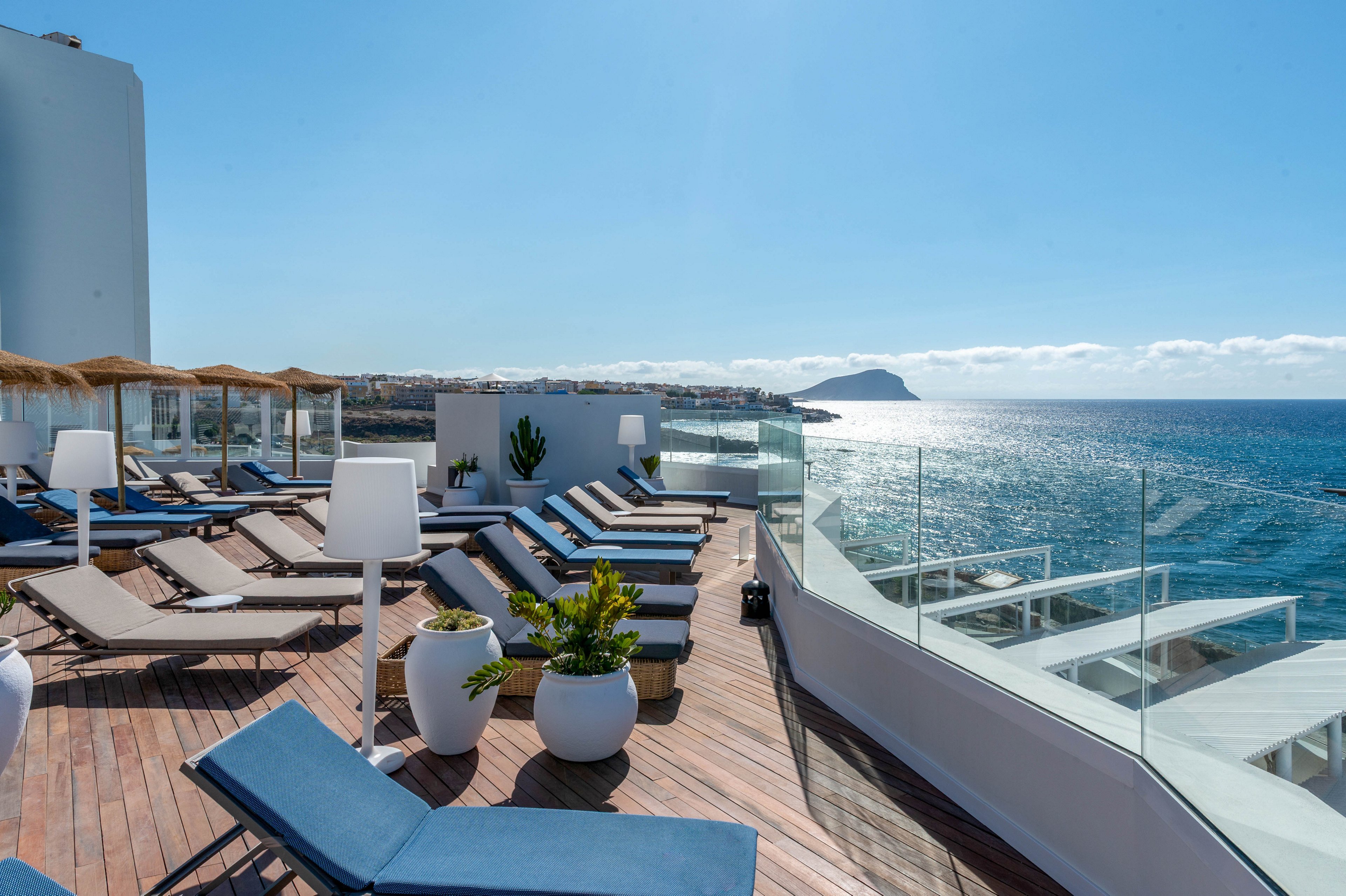 A modern hotel terrace overlooks a bright blue sea under a clear sky. Numerous sun loungers with cushions are arranged on a wooden deck beside white planters and contemporary lamps. Glass railings frame the edge of the terrace, providing an unobstructed ocean view. In the distance, coastal buildings and a small island or headland are visible along the horizon.