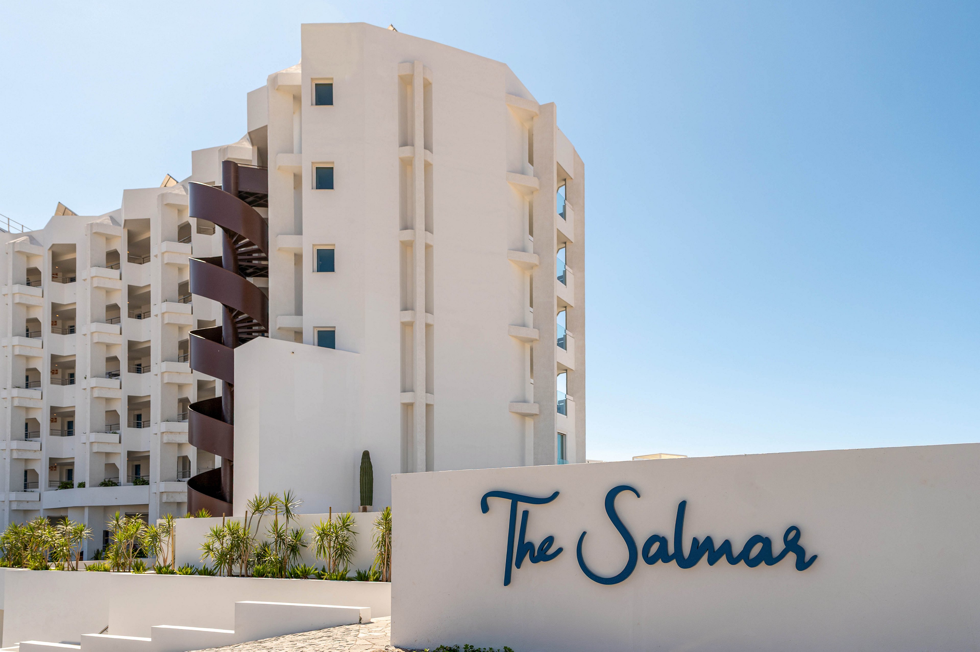 A modern white hotel building stands under a clear blue sky, with multiple balconies and an external spiral staircase visible. In the foreground, a white entrance wall prominently displays the name "The Salmar" in blue script. Low, landscaped greenery and palm-like plants line the front of the property, enhancing the coastal resort atmosphere. The bright sunlight and clean architectural lines create a fresh, inviting vacation setting.