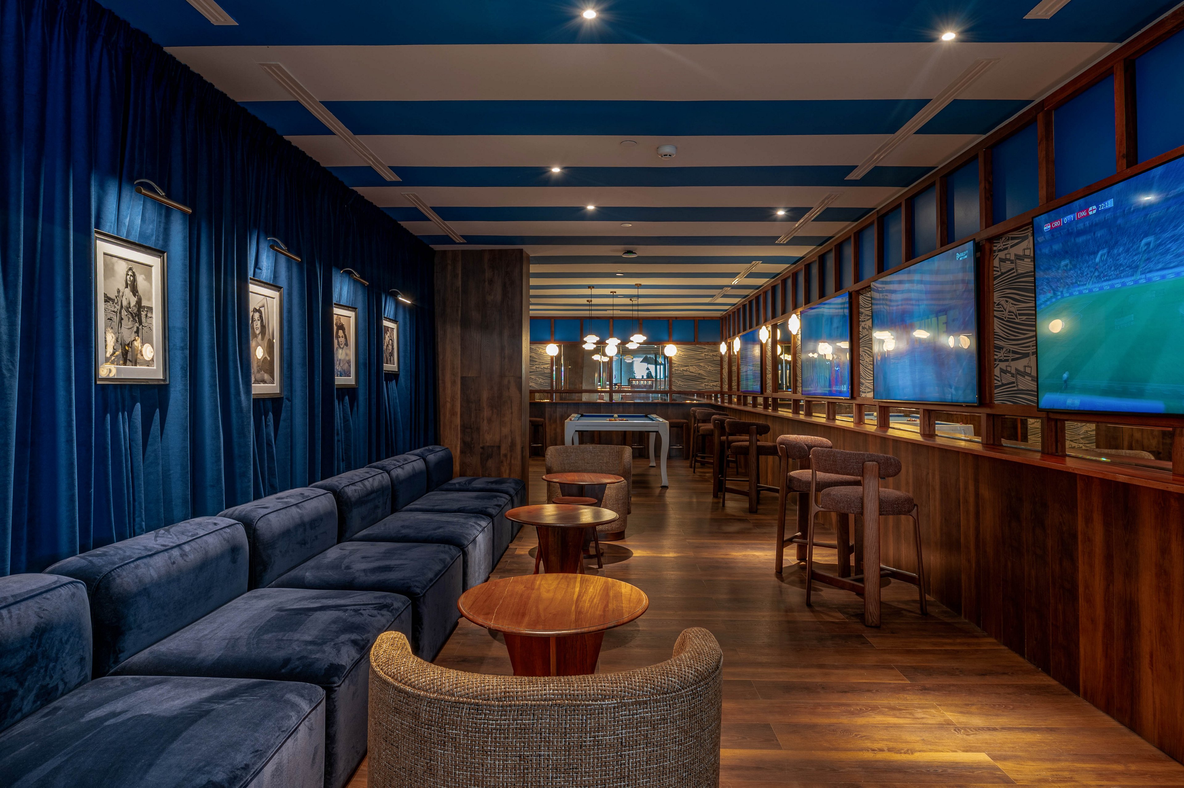 A contemporary lounge bar interior features plush blue velvet sofas along one wall and high wooden stools opposite. Warm wooden flooring and paneling contrast with deep blue curtains and ceiling accents. Framed black-and-white artwork decorates the walls, while large screens display a sports match. Soft ambient lighting and a mix of low tables and bar-height seating create a relaxed, upscale atmosphere.