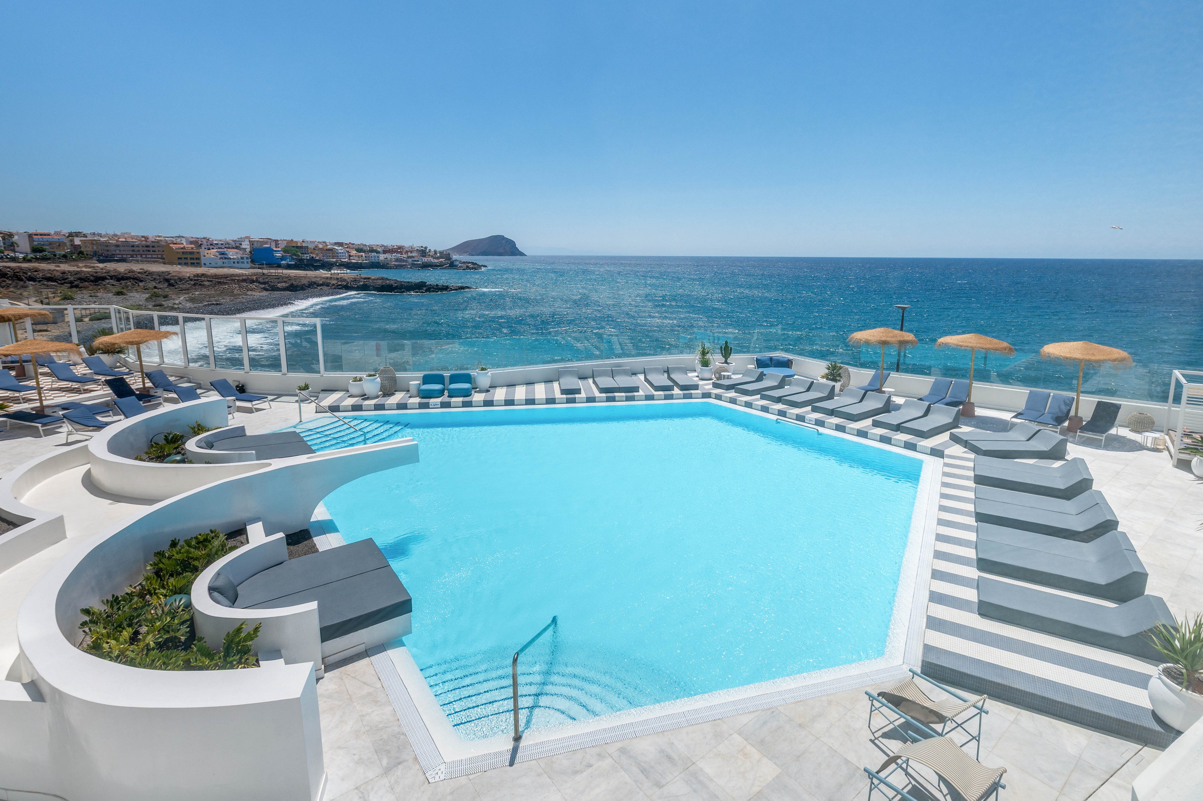 A contemporary hotel swimming pool overlooks a calm blue ocean under a clear sky. Curved white architecture surrounds the pool, with numerous sun loungers arranged in neat rows. Yellow parasols line the edge of the terrace, and glass railings frame the view toward distant coastal buildings and a rocky headland. The bright, minimalist design emphasizes relaxation and resort-style luxury by the sea.