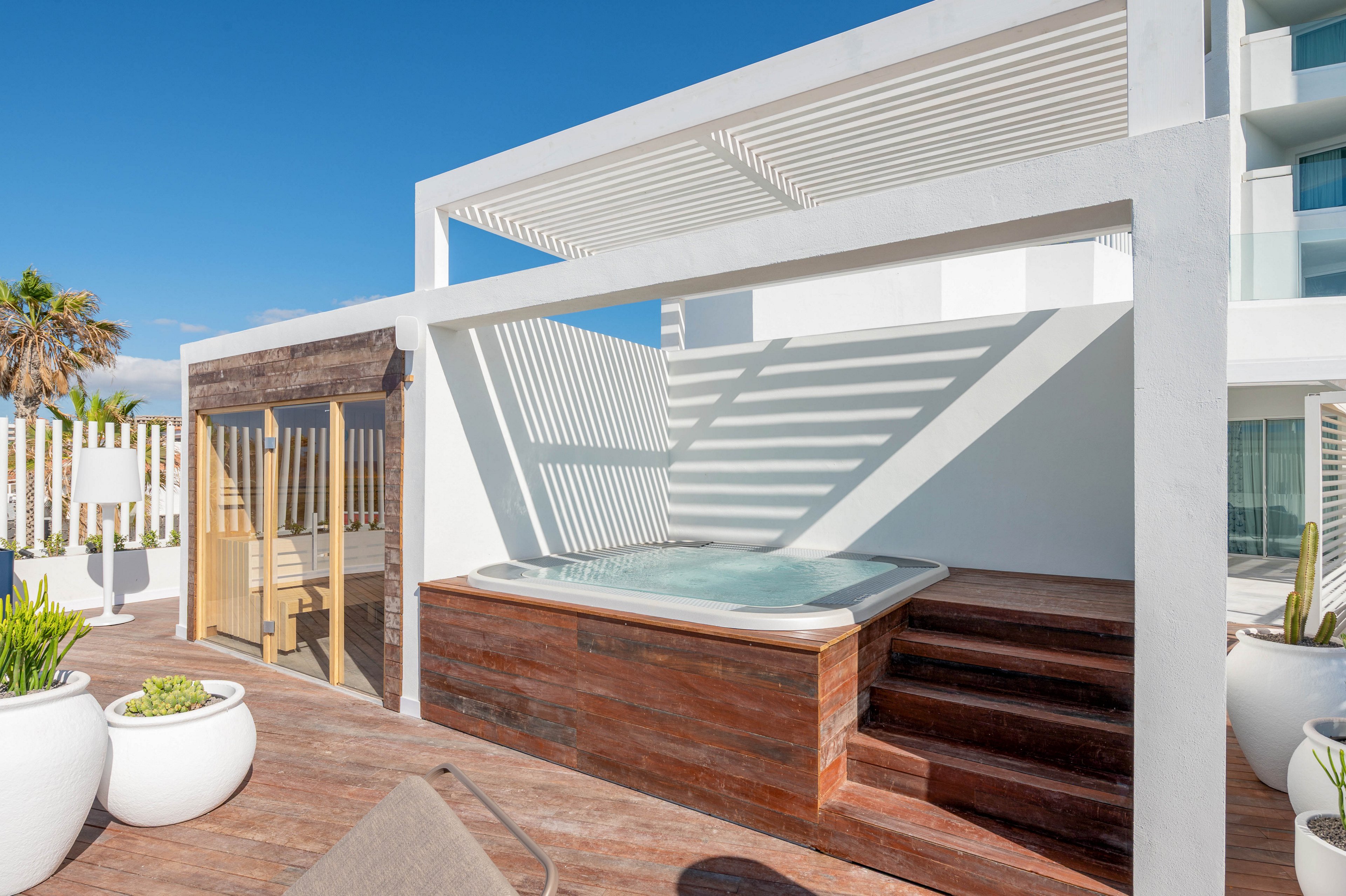 A contemporary rooftop terrace features a raised jacuzzi framed by a white pergola casting linear shadows. Wooden steps lead up to the hot tub, which is clad in warm wood panels contrasting with the white architecture. To the side, a small wooden sauna structure stands near large white planters with green plants. The bright outdoor setting suggests a relaxing spa environment under clear blue skies.
