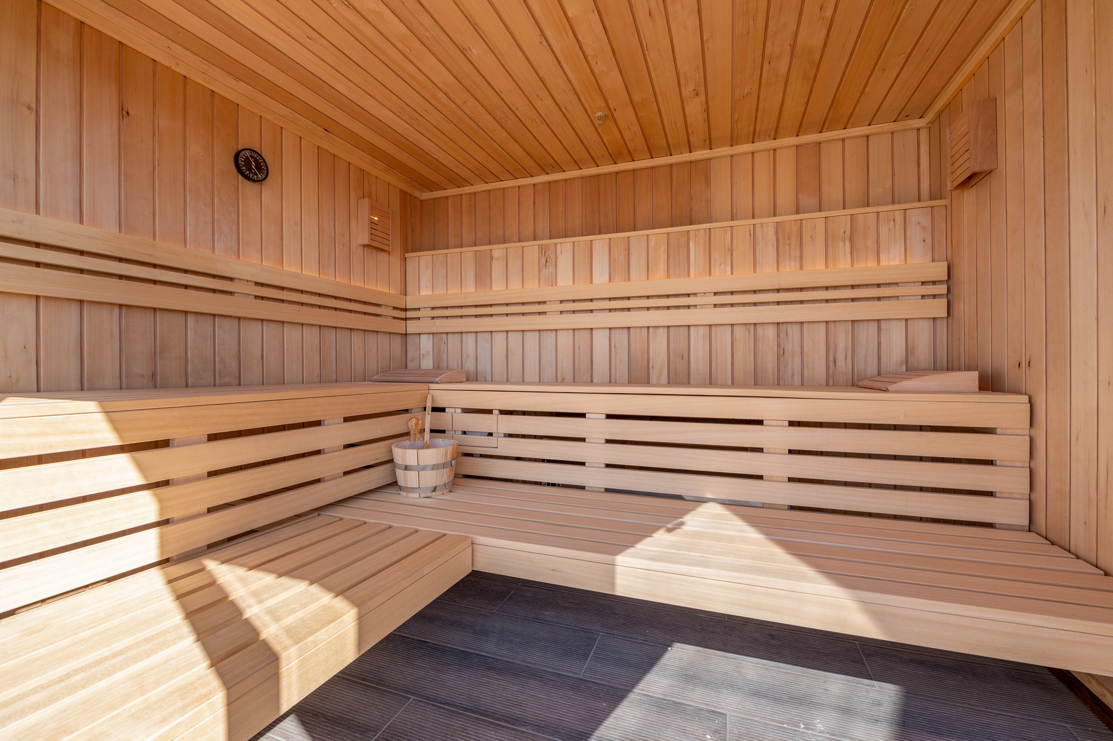 Image of a bright, modern wooden sauna interior featuring tiered benches. The space is constructed from light-toned wood with horizontal slats and clean, minimal lines. A wooden bucket and ladle sit on one of the benches, suggesting preparation for a sauna session. The floor is finished in dark tiles, creating contrast with the warm wood surfaces.