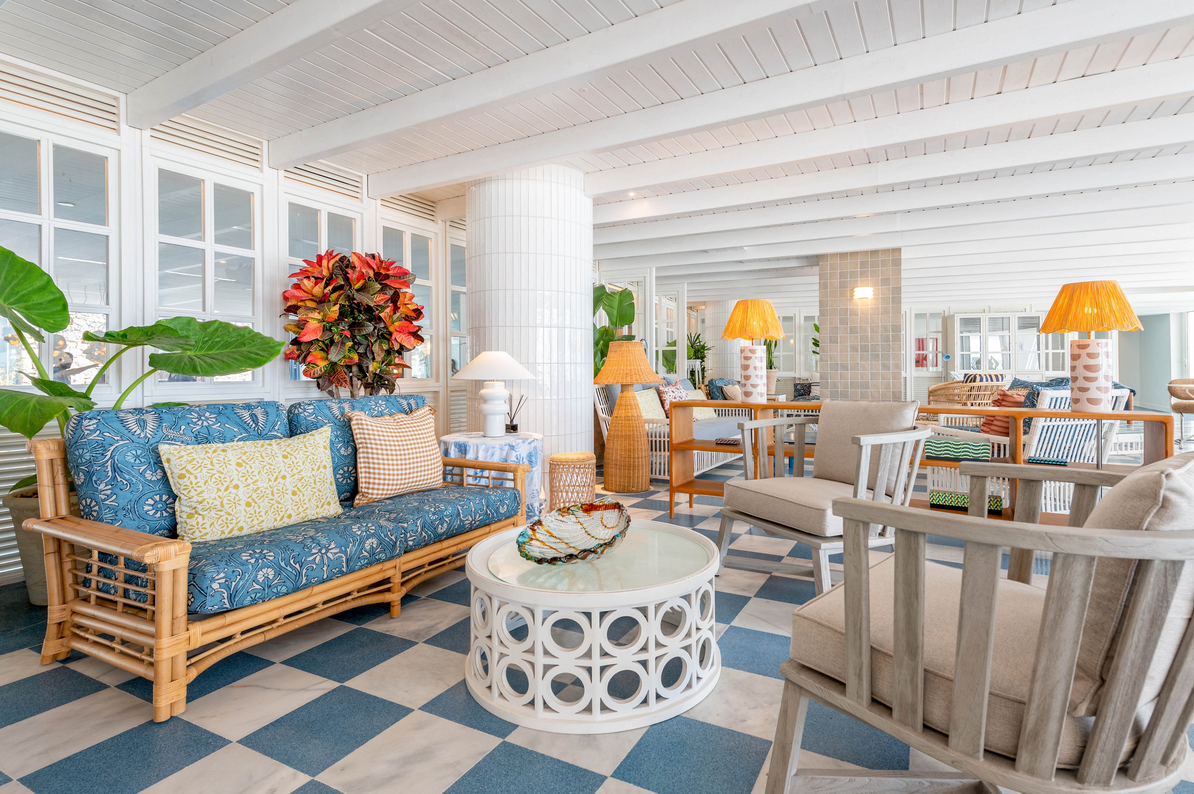 A spacious indoor lounge features rattan and wooden seating with patterned cushions arranged around a circular coffee table. The room has a light, airy feel with white paneled ceilings and large windows in the background. Blue and white checkered flooring adds a coastal touch, complemented by warm table lamps and decorative plants. The overall setting suggests a relaxed, beach-inspired social or waiting area.