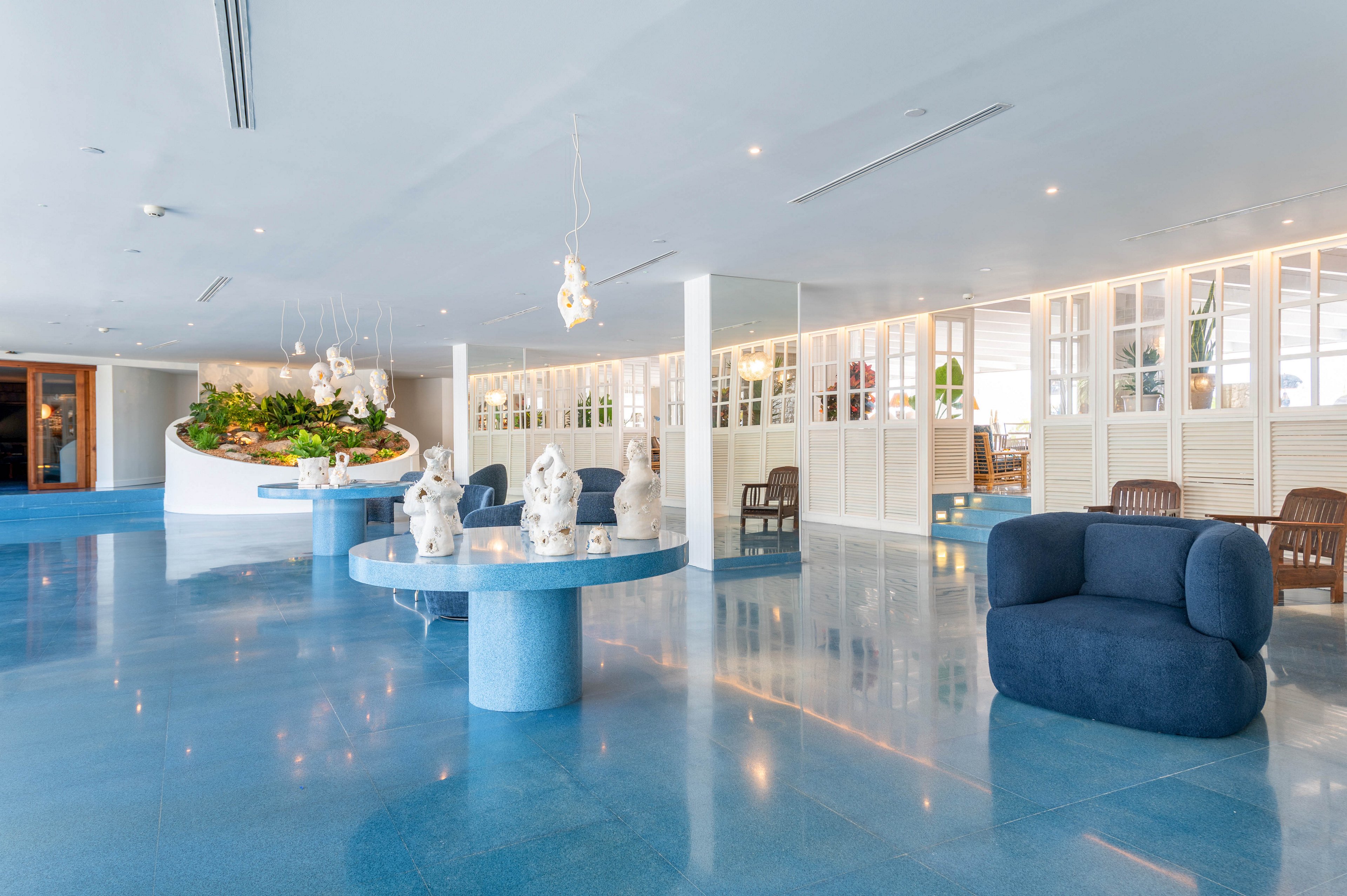Spacious contemporary lobby interior features glossy blue flooring and rounded blue lounge furniture. A central circular table displays white sculptural art pieces under hanging pendant lights. In the background, white paneled walls and large windows create an airy, bright atmosphere. A curved planter filled with lush green plants adds a natural accent to the clean, minimalist design.