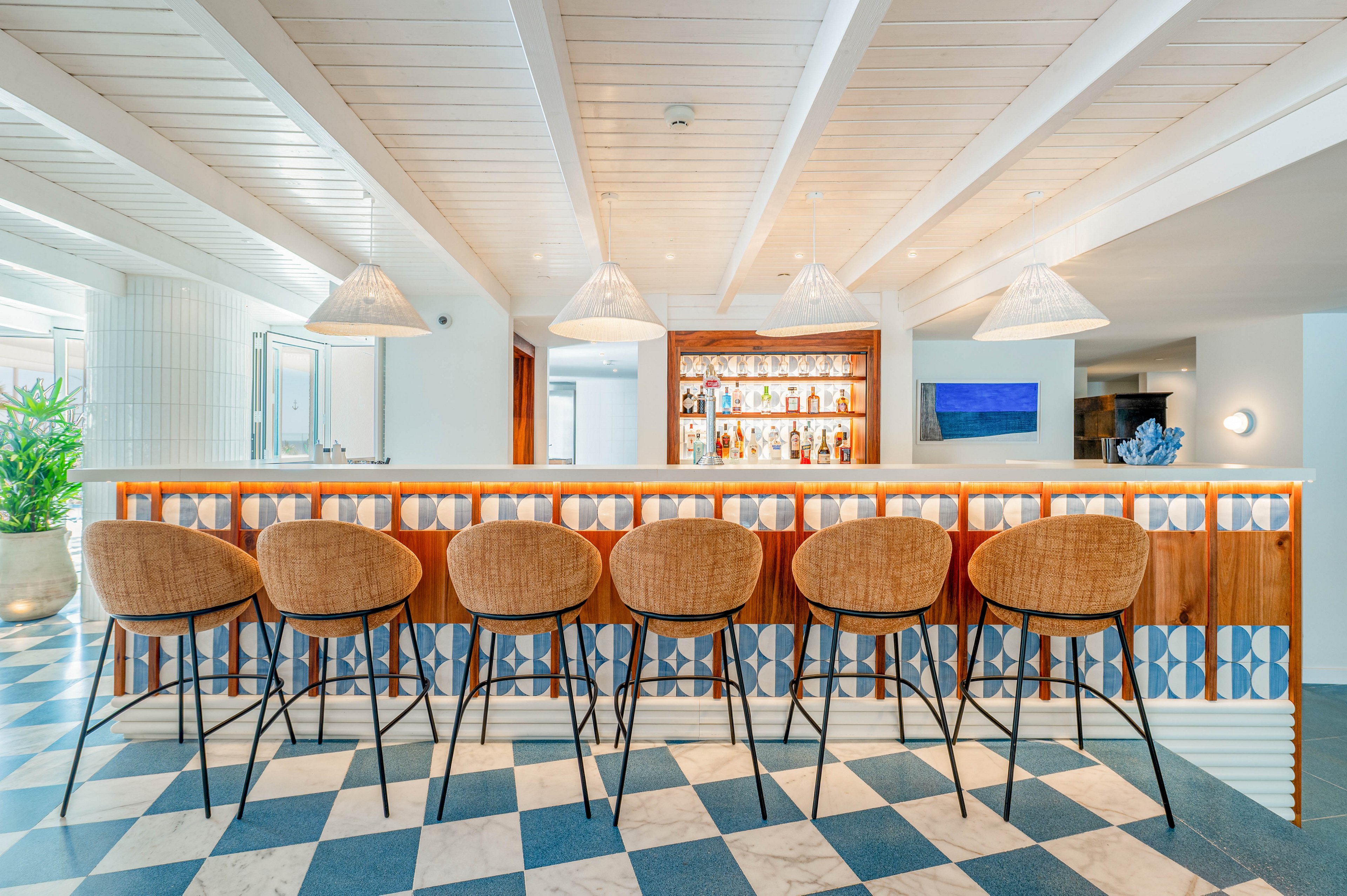 A stylish bar interior features a row of wicker bar stools lined up along a patterned tiled counter. The space is bright and airy, with white wooden ceiling beams and large windows letting in natural light. Bottles and glassware are neatly arranged on shelves behind the bar, adding visual interest. Blue and white tones in the floor tiles and wall art create a fresh, coastal atmosphere.